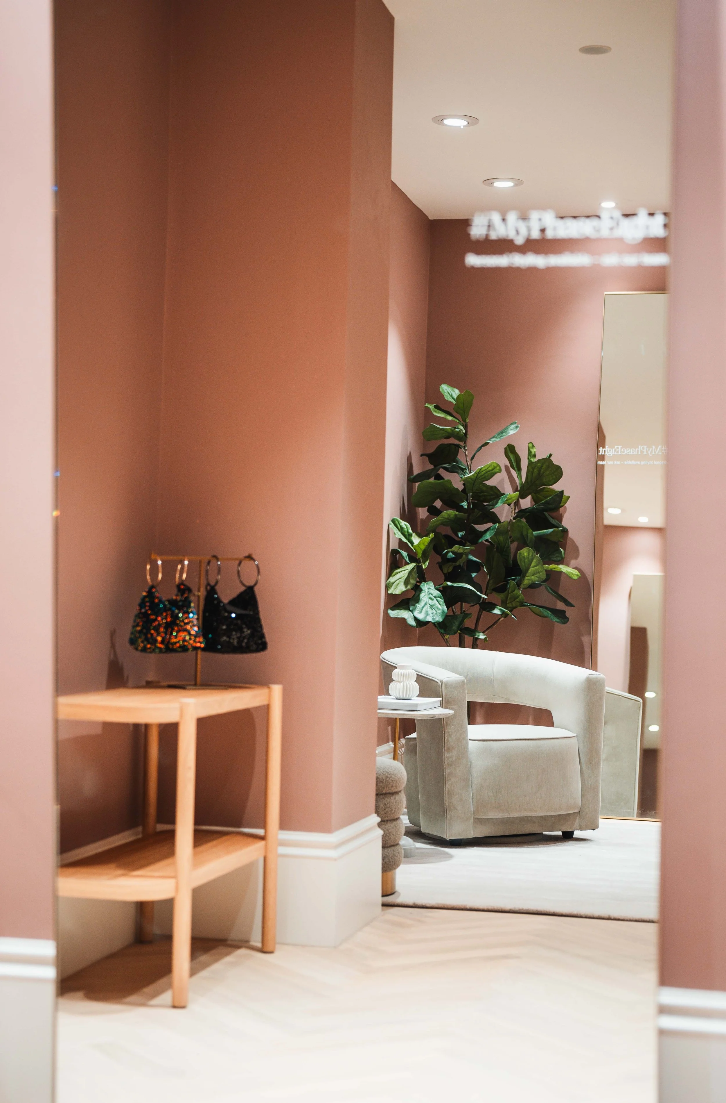 A boutique store interior with a large mirror reflecting a pink wall, a beige armchair, a small side table with decorative items, a leafy potted plant, and colorful handbags on a stand.