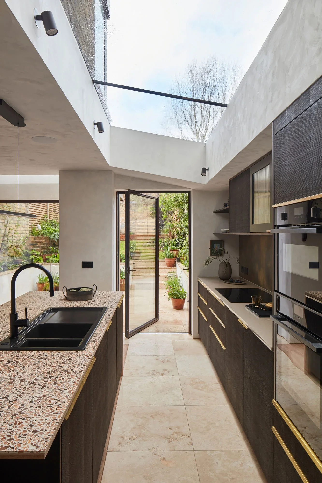 Modern kitchen with a skylight, open door leading to an outdoor garden with potted plants, dark cabinetry with gold handles, granite countertop, black sink, and black fixtures, beige tiled floor.