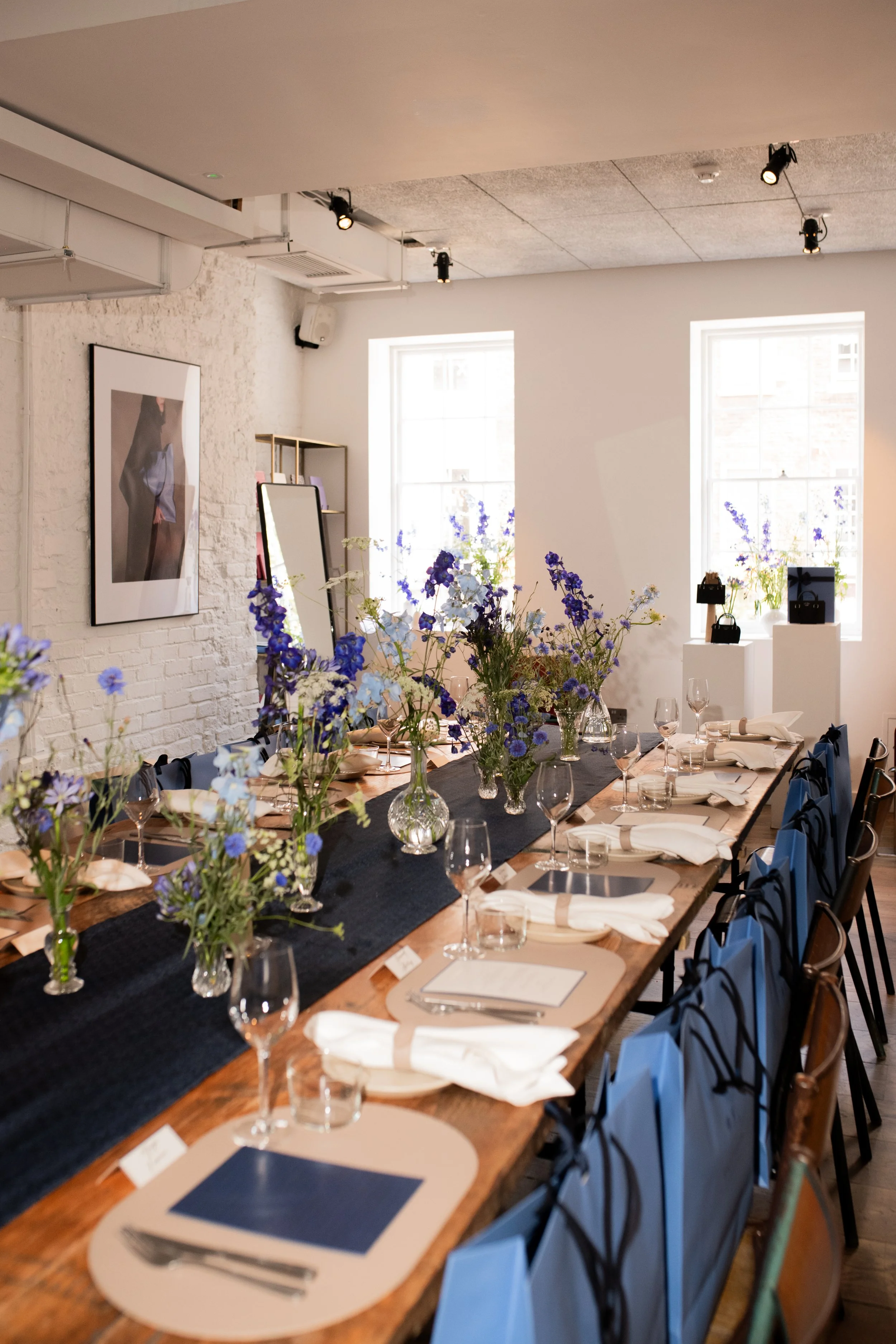 A long wooden table set for a formal dinner with white napkins, glassware, and floral centerpieces in vases. The room has white brick walls, two large windows with purple flowers on the windowsills, and modern art on the walls.