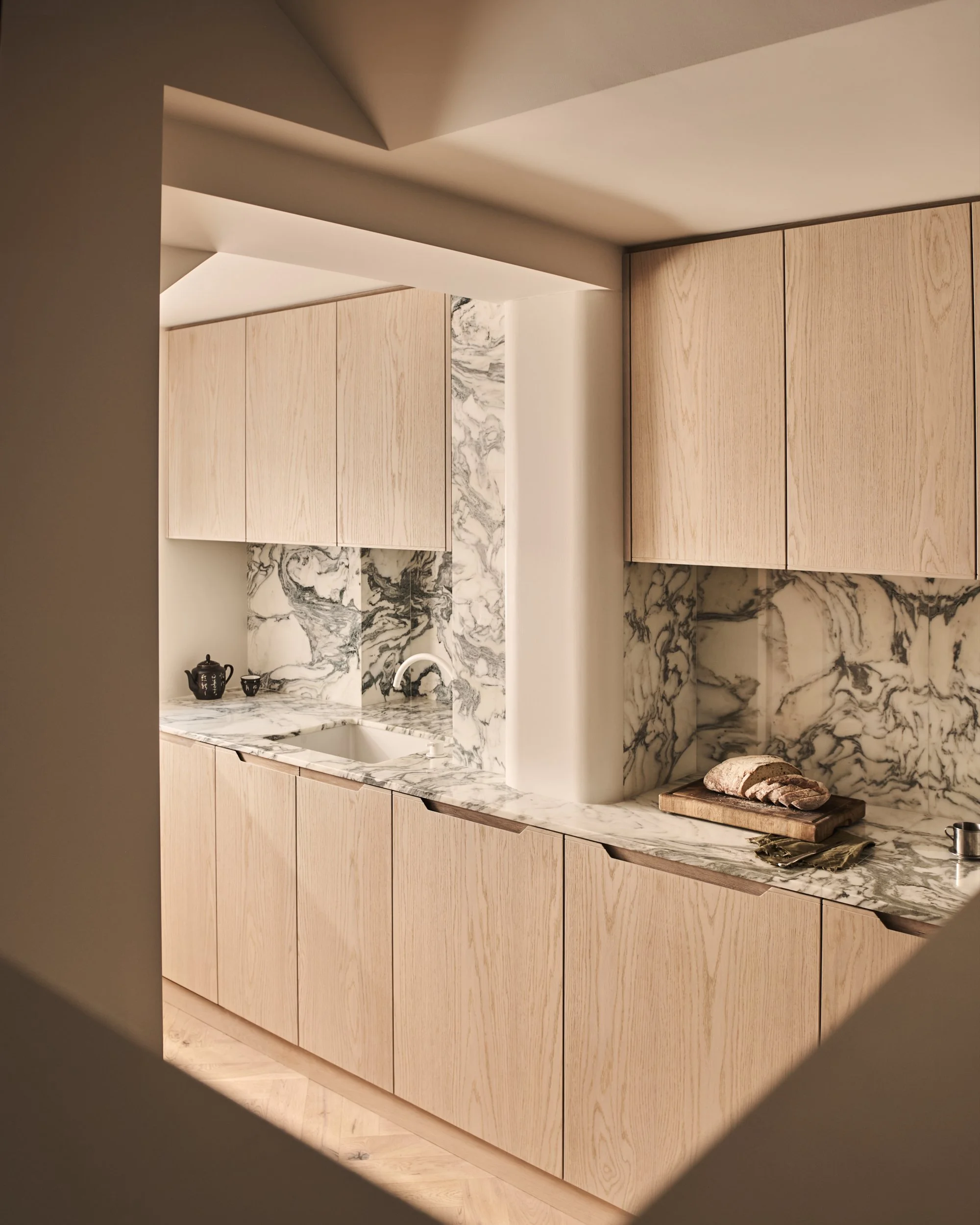 Modern kitchen with light wood cabinets, marble backsplash, and countertop, with a loaf of bread on a cutting board.