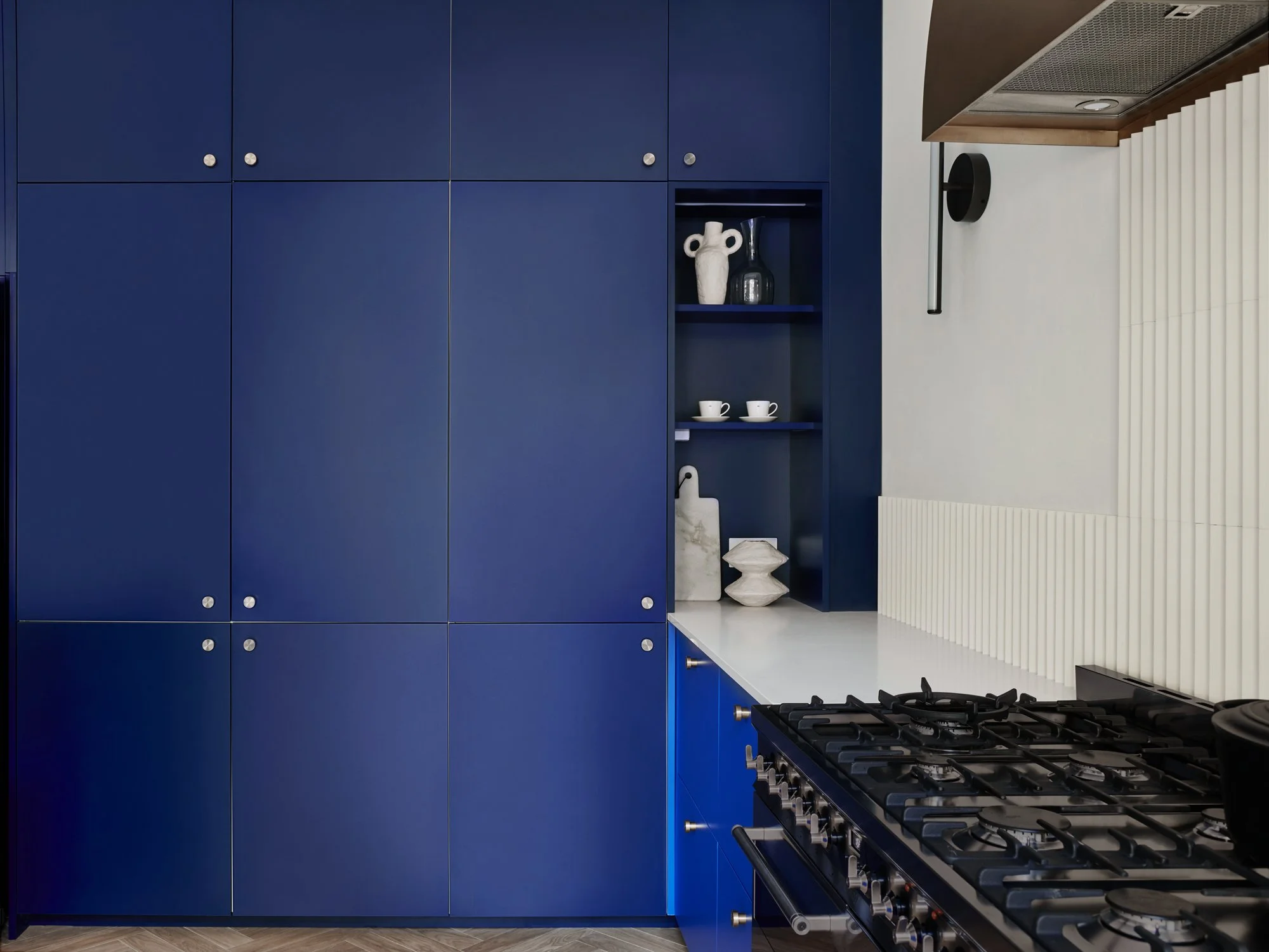 Modern kitchen with blue cabinetry, white countertop, and stainless steel gas stove. Open shelving displays white vases, cups, and decorative objects. The wall has a white textured panel and a vent hood above the stove.
