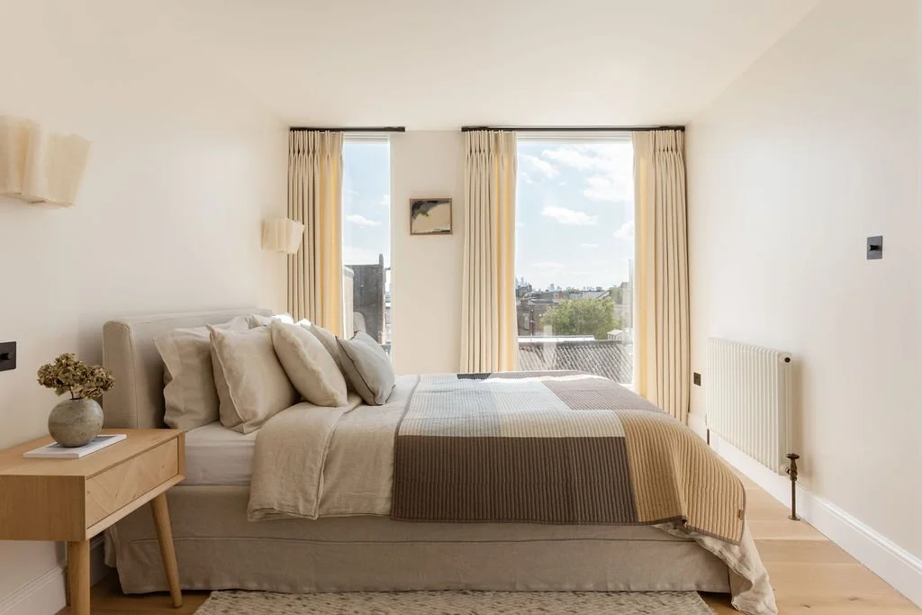 Bright and minimalistic bedroom with a bed, decorated in neutral tones, facing large windows with cream curtains, overlooking cityscape and blue sky.