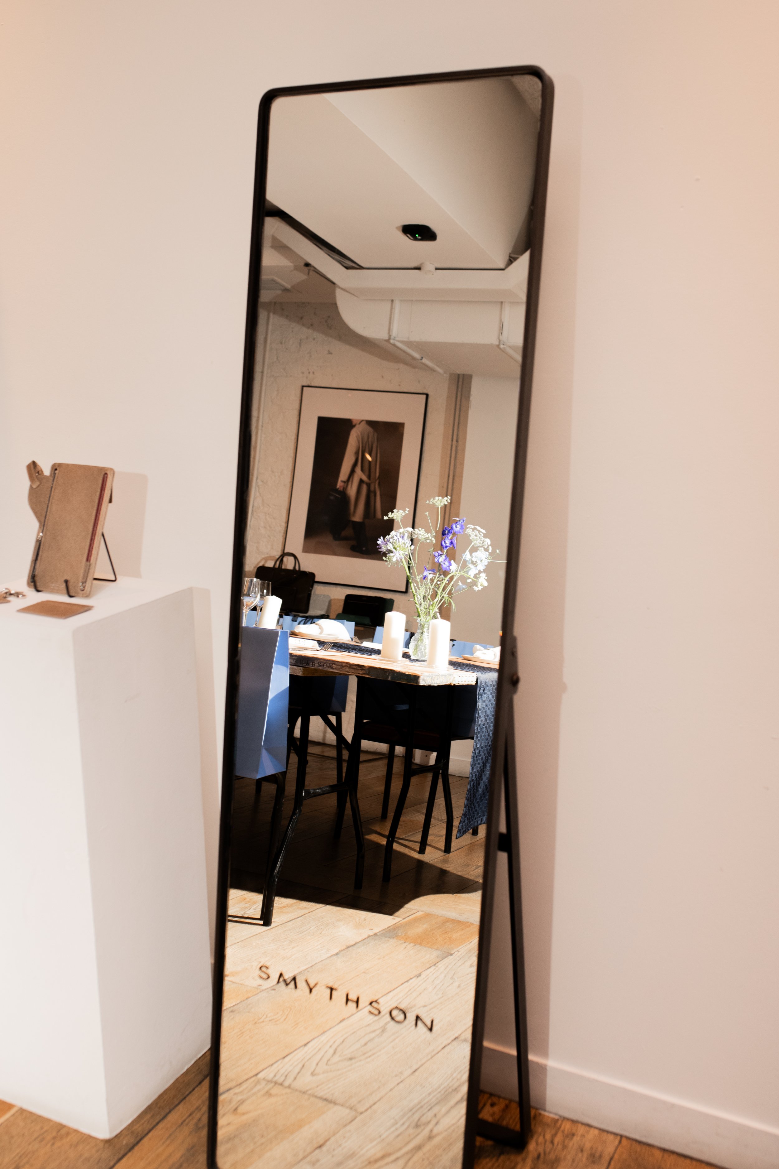 A tall, rectangular mirror reflecting a dining area with a table, chairs, and a floral centerpiece. The mirror has the word "SMYTHSON" on the wooden floor in front of it.