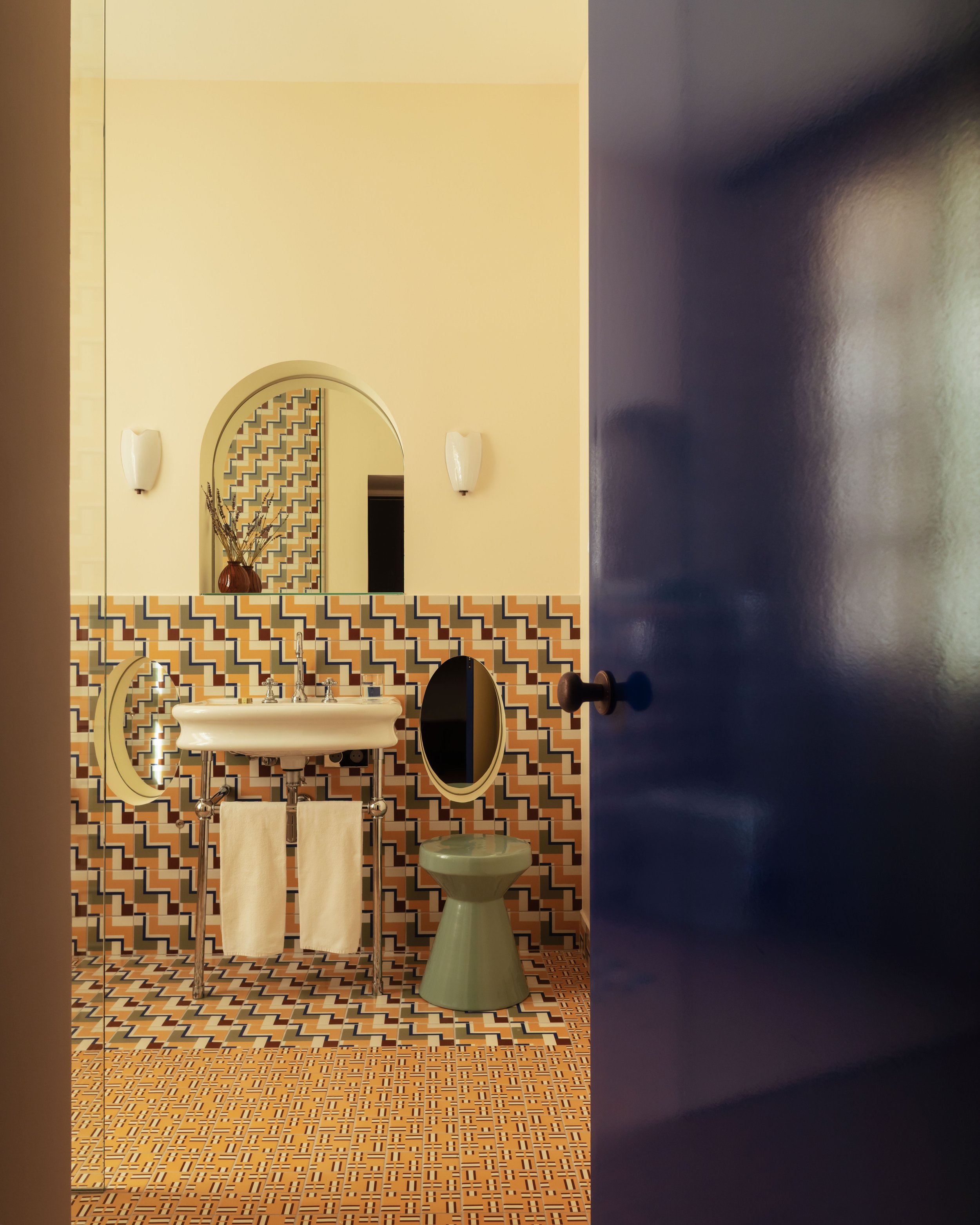 View of a vintage bathroom with a patterned tile wall and floor, a small sink with two towel racks, a green toilet, and a mirror on the wall. The door is slightly open.