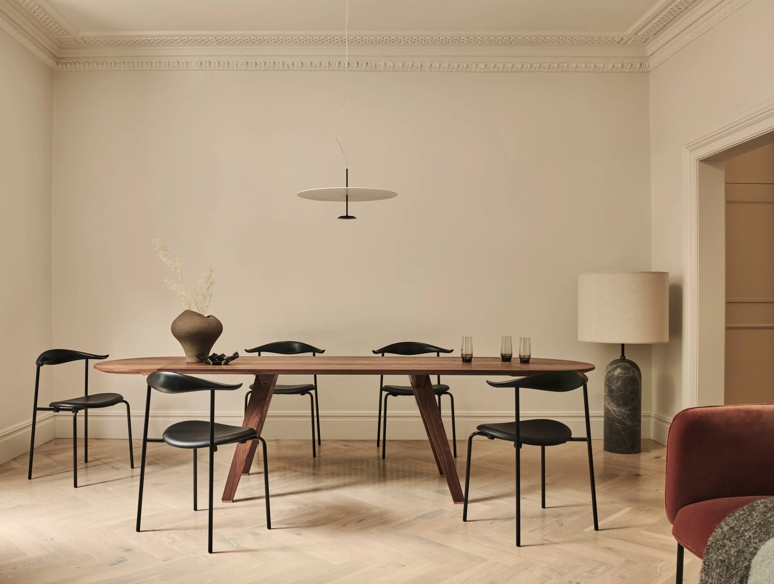 Minimalist dining room with a wooden table, five black chairs, a large beige vase with dried flowers, a modern pendant light, and a tall floor lamp with a beige shade in a corner.