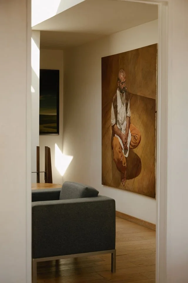 Interior of a home featuring a large painting of a seated monk or spiritual figure, a gray couch, a wooden dining table, and chairs, with a natural wood floor.