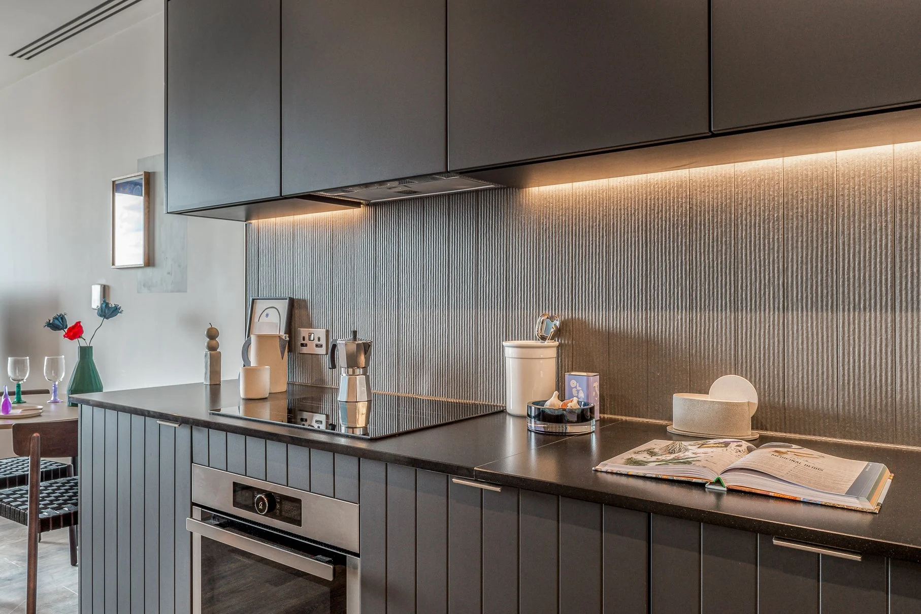 Modern kitchen with dark cabinets, a black countertop, and textured backsplash. Items on the counter include a book, a bowl, and containers. To the left, a dining table with a vase and flowers is visible.