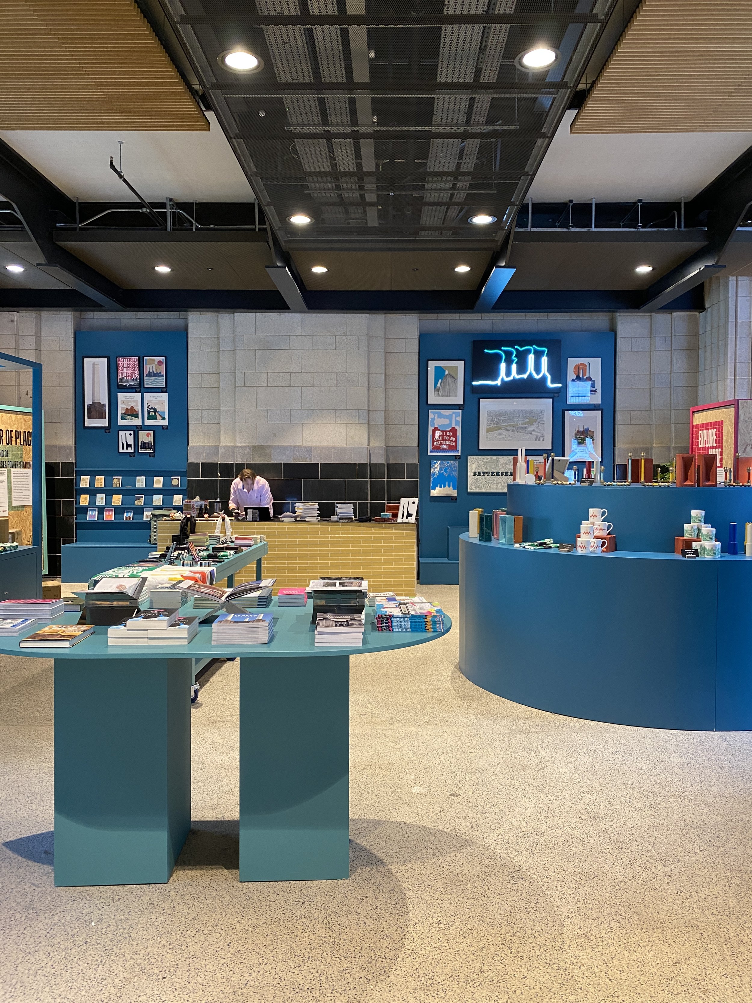 Interior of a modern bookstore or gift shop with a blue display counter, books, and merchandise on tables, and artwork and neon sign on the blue wall behind the counter.