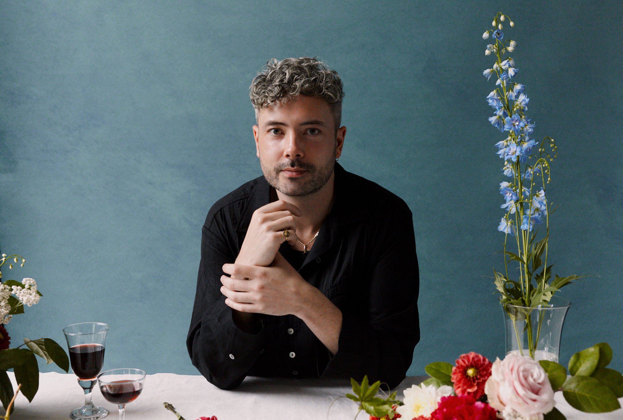 A man with curly gray hair and a beard sits at a table decorated with flowers and glasses of red wine, against a teal background.