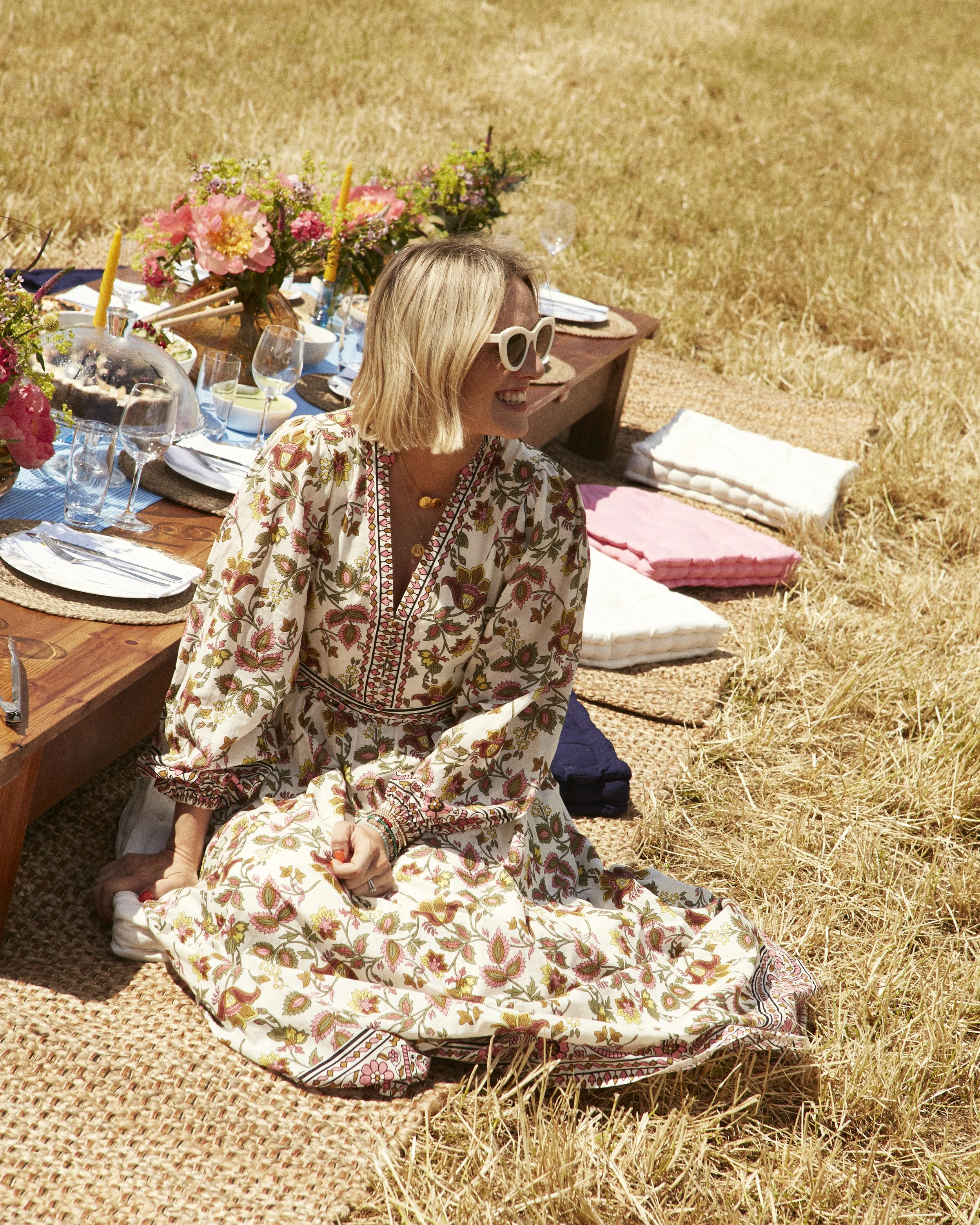 A woman in a floral dress with large white sunglasses sitting on a woven mat at a picnic table outdoors on dry grass, with colorful flowers, plates, glasses, and candles on the table.