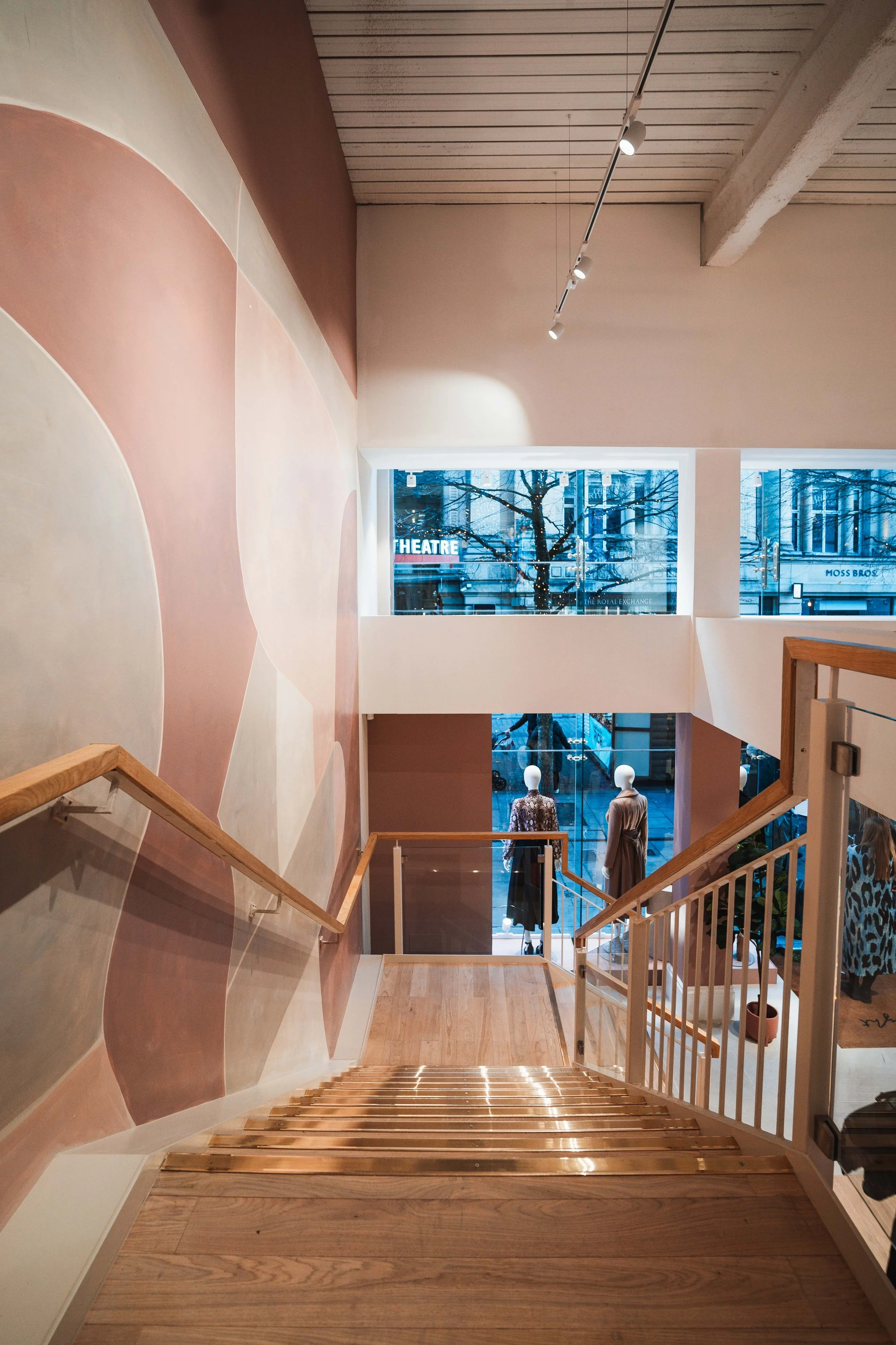 Interior view of a modern store with staircase, mannequins dressed in clothing near large windows, and city street outside.