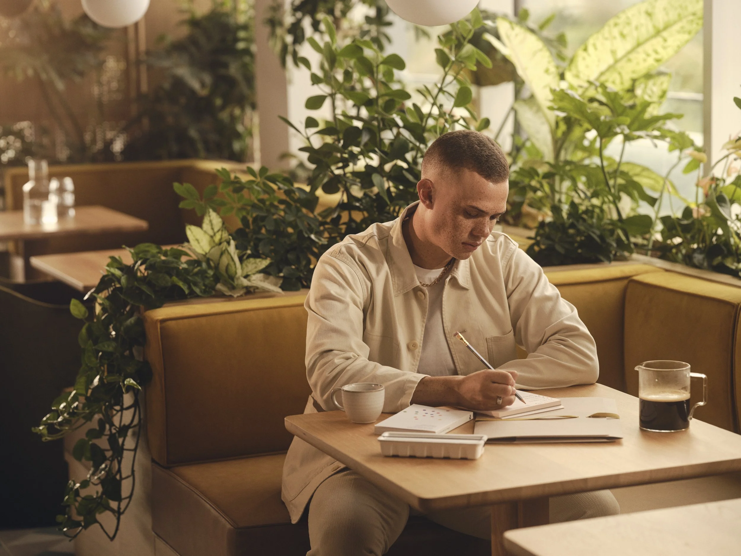 A young man with short hair and freckles sitting in a cozy cafe, writing in a notebook on a wooden table with a coffee cup and a glass of soda nearby, surrounded by green plants and soft lighting.