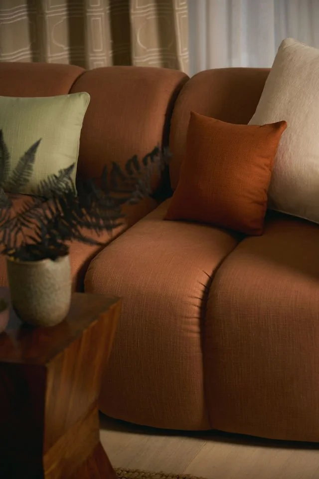Close-up of a brown sofa with three decorative pillows in green, rust, and beige, next to a wooden side table with a potted plant, in a room with patterned curtains in background.