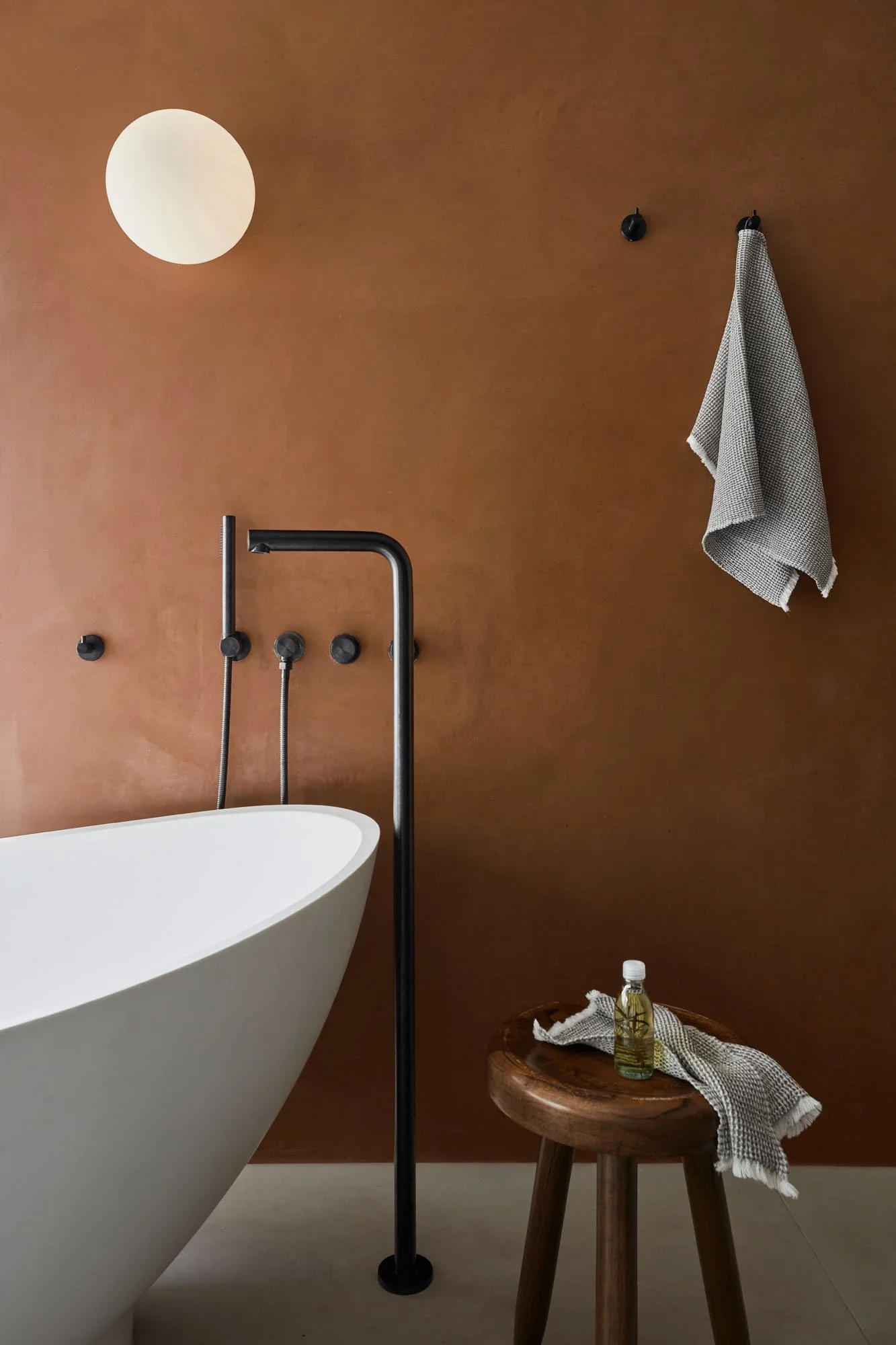 Modern bathroom with a white bathtub, brown wall, black fixtures, towel hanging on wall, small wooden stool with a bottle of soap and a washcloth, and a wall-mounted round mirror.