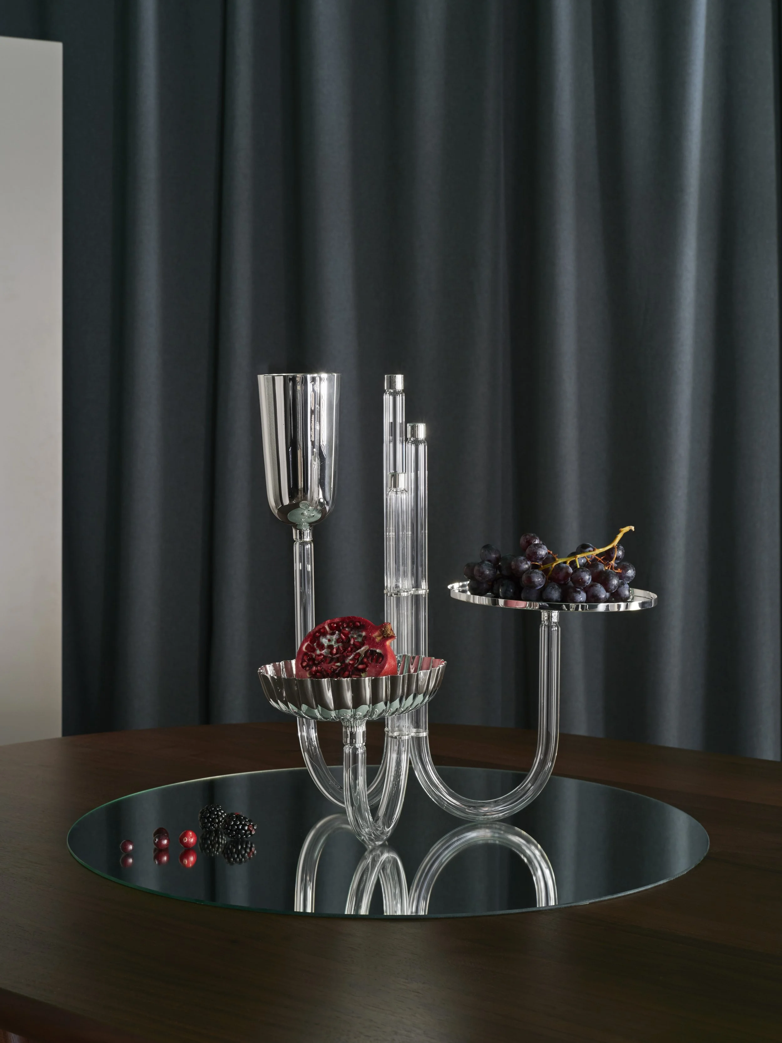 Elegant glass fruit holder with grapes, pomegranate, and berries on a wooden table with a round mirror surface.