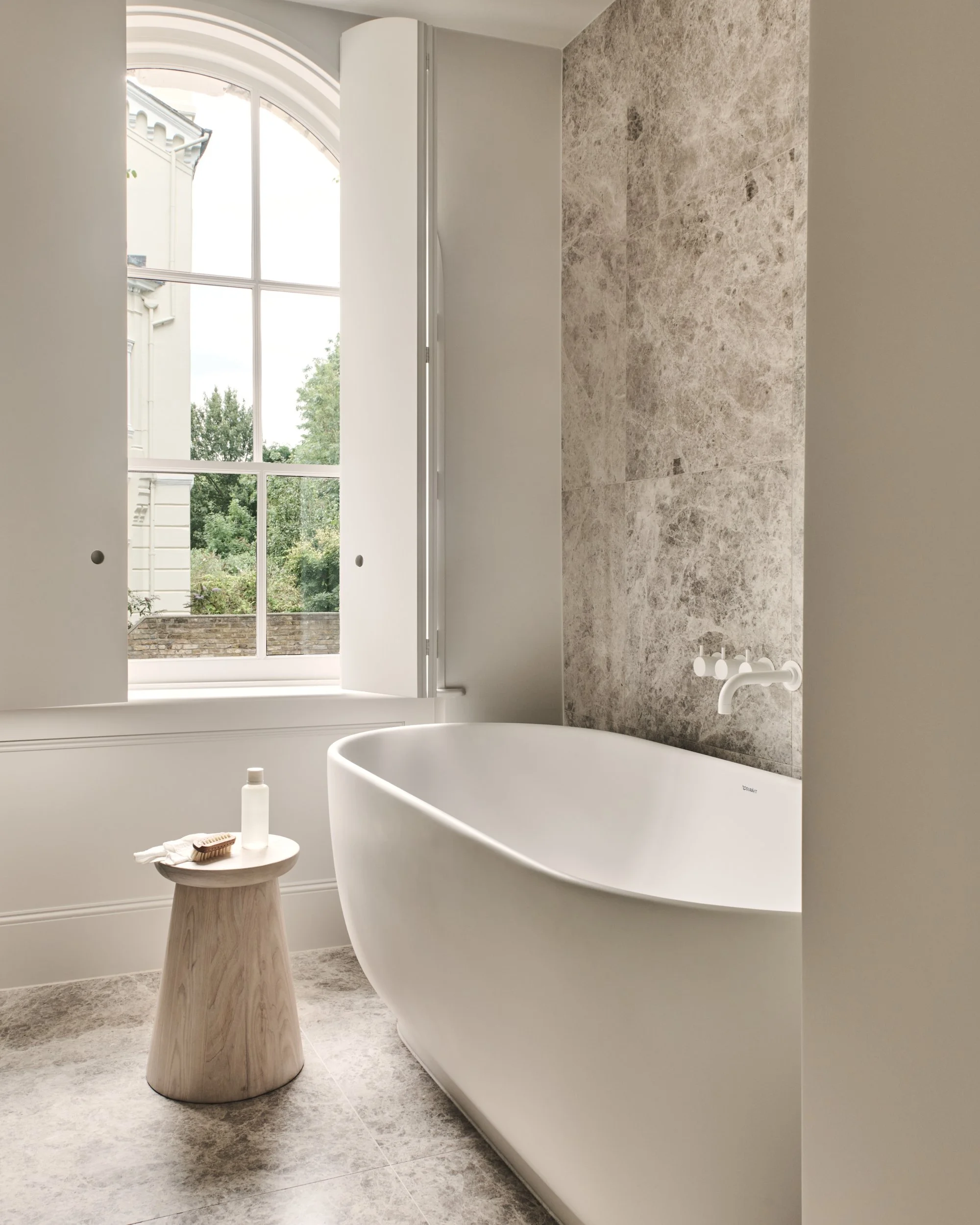 Modern bathroom with a white oval bathtub, a small wooden stool with toiletries, a large window with white shutters, and a textured stone wall.