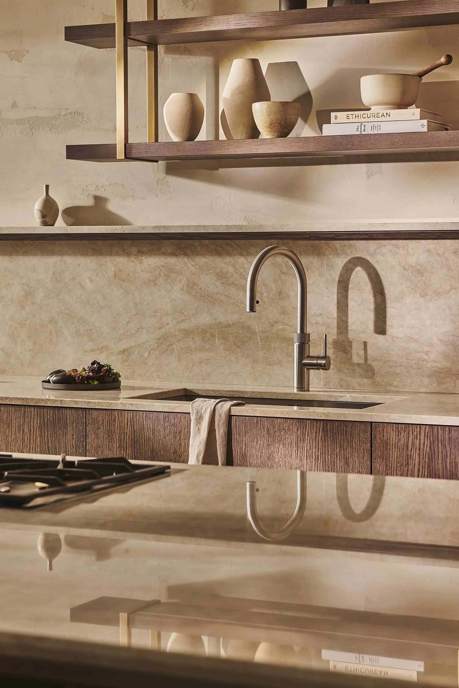 A modern kitchen with beige stone countertops, wooden cabinets, and a stainless steel faucet above a sink. Open wooden shelves above display beige ceramic vases and a stack of books, including one titled 'ETHICUREAN.' The reflection of the faucet and