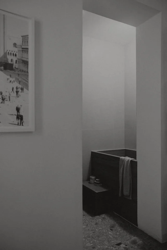 Black and white photo of a corner in a room with a small wooden soaking tub with a towel hanging on the side and some containers on a small table nearby. Part of a framed picture is visible on the wall to the left.