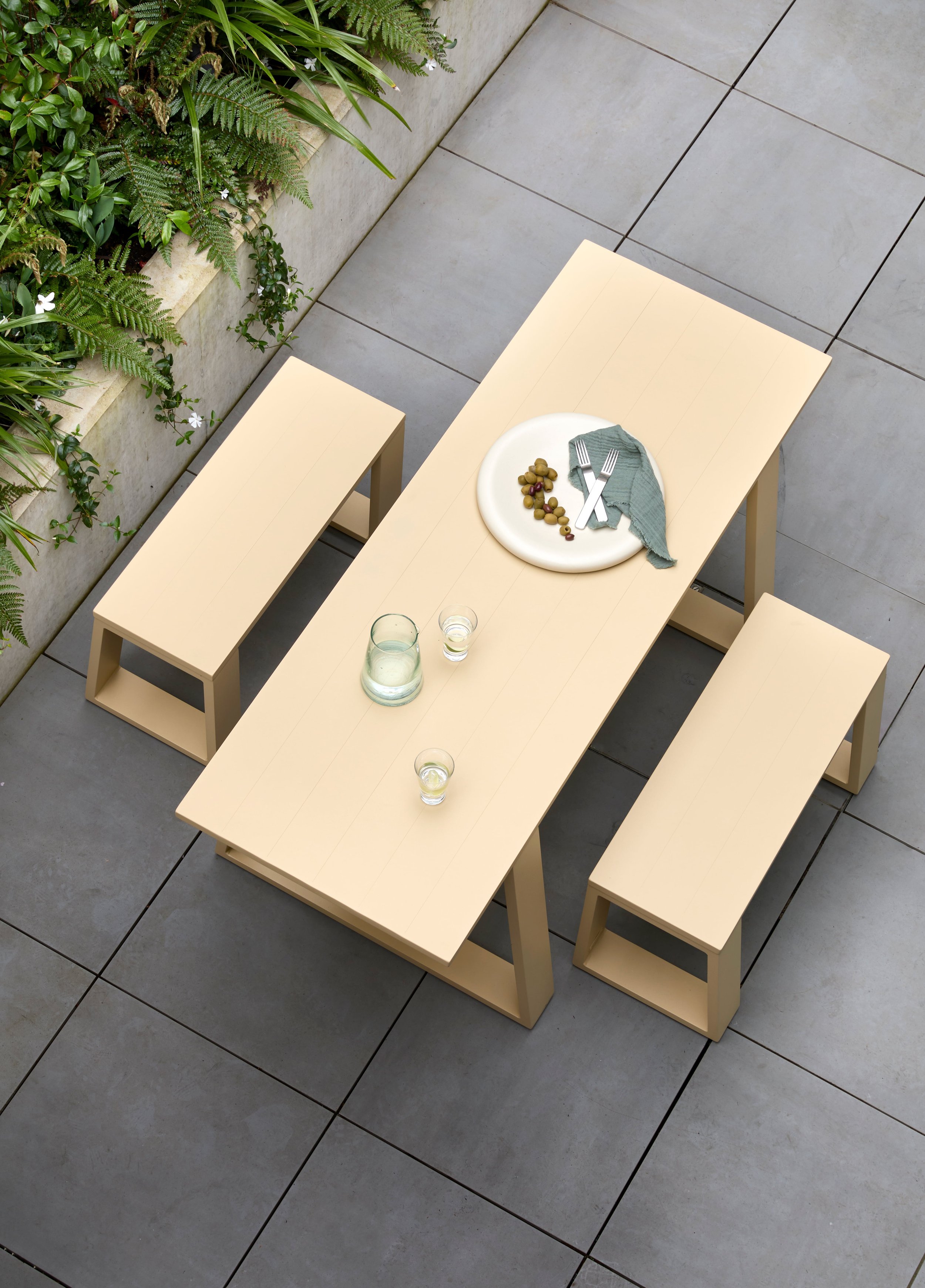 A beige outdoor dining table set on a tiled patio with three matching benches. The table has a white plate with olives, a blue cloth napkin, a pitcher of water, and three glasses with drinks.
