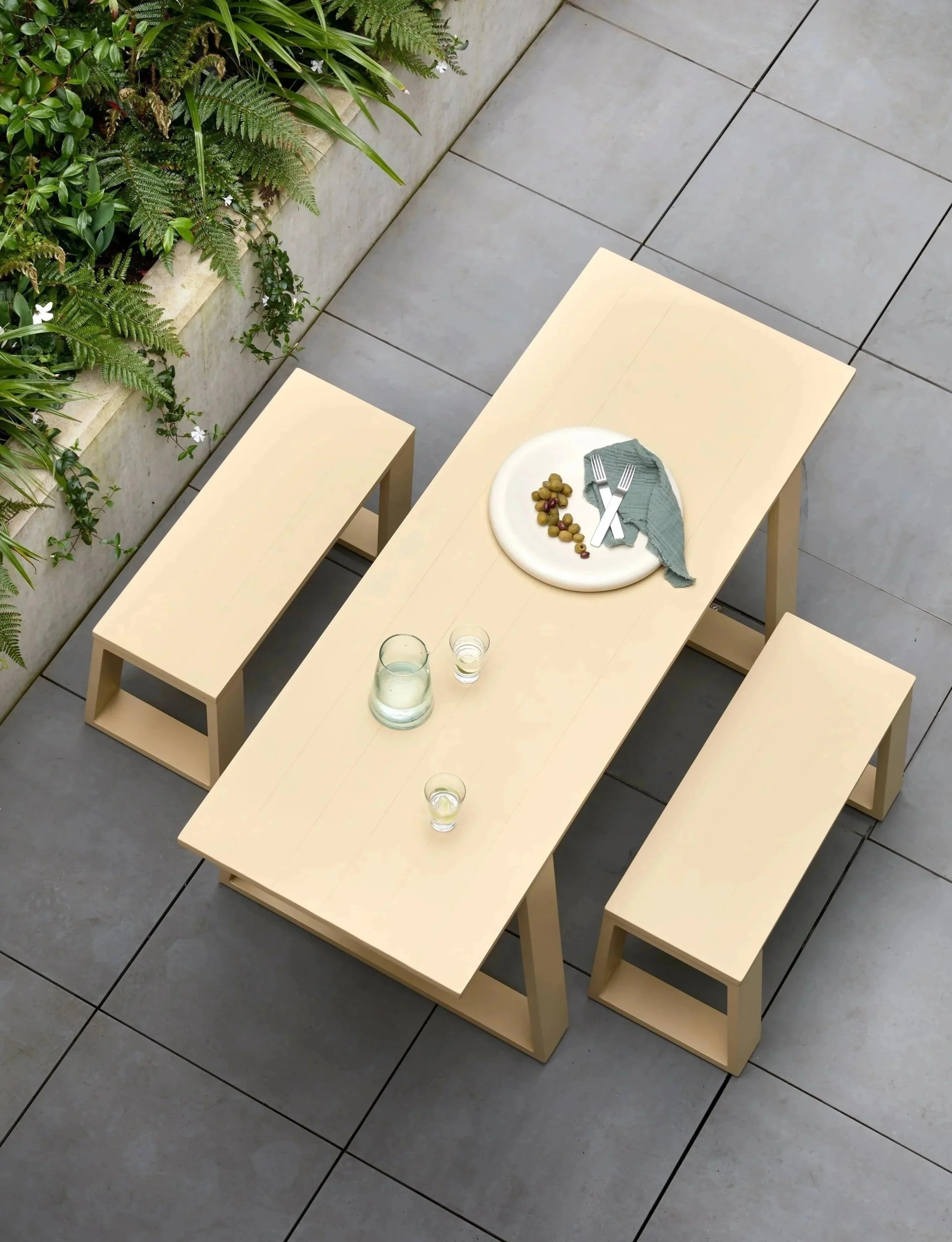 A minimalist outdoor dining table with three matching benches, set on a tiled patio adjacent to green plants; a white plate with grapes, a gray napkin, and cutlery, along with a glass pitcher and glasses of water.