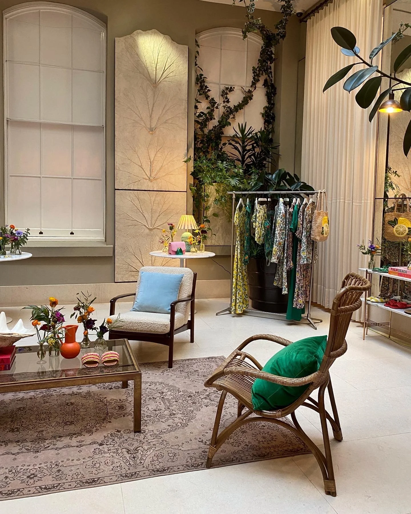 Interior of a cozy boutique with vintage and bohemian decor, displaying a clothing rack, armchairs, floral arrangements, and decorative wall panels.
