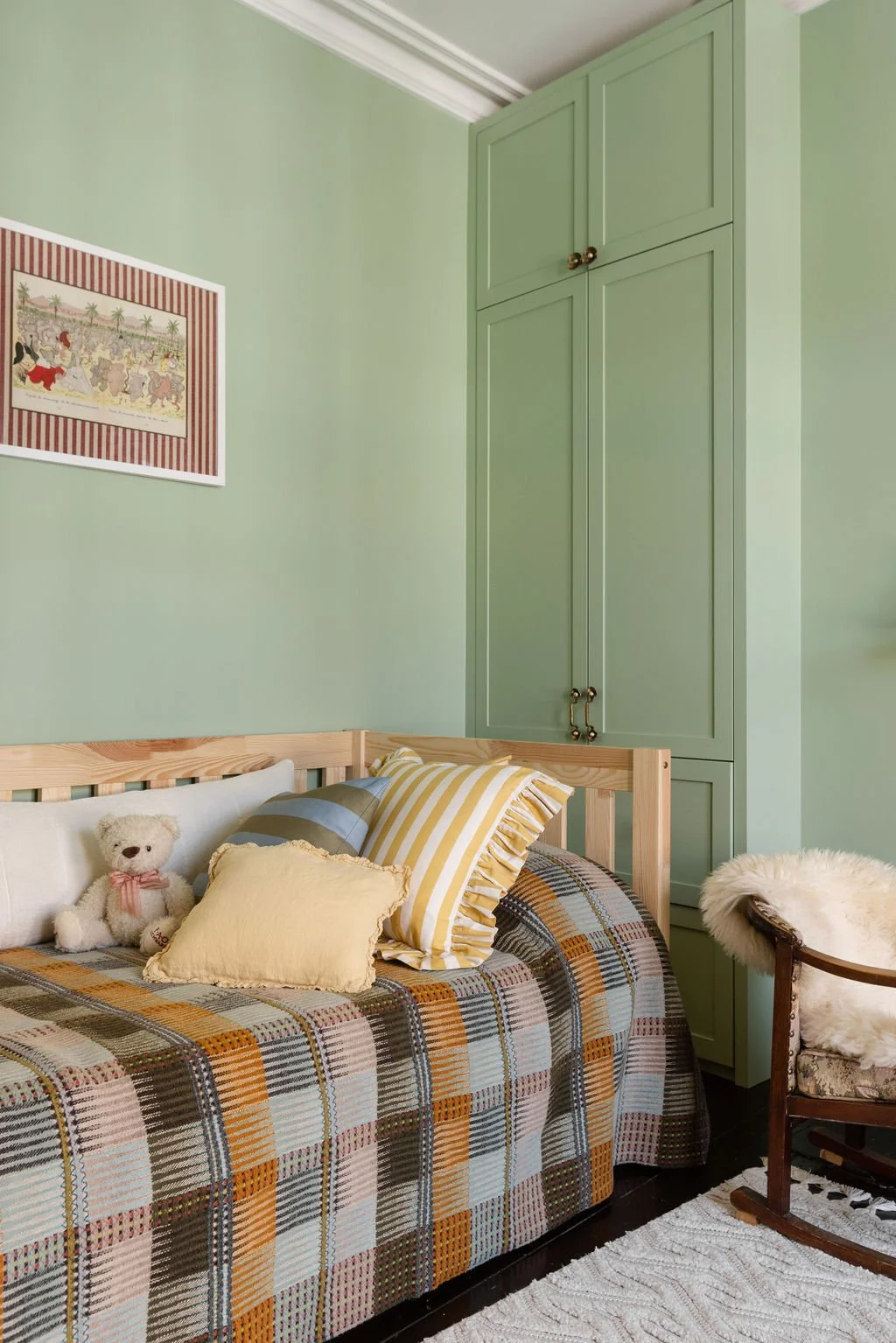 A cozy bedroom corner with a bed that has a plaid bedspread and various pillows, including a yellow ruffled pillow and a striped blue and yellow pillow. There is a teddy bear on the bed. To the right, a vintage wooden rocking chair with white furry p