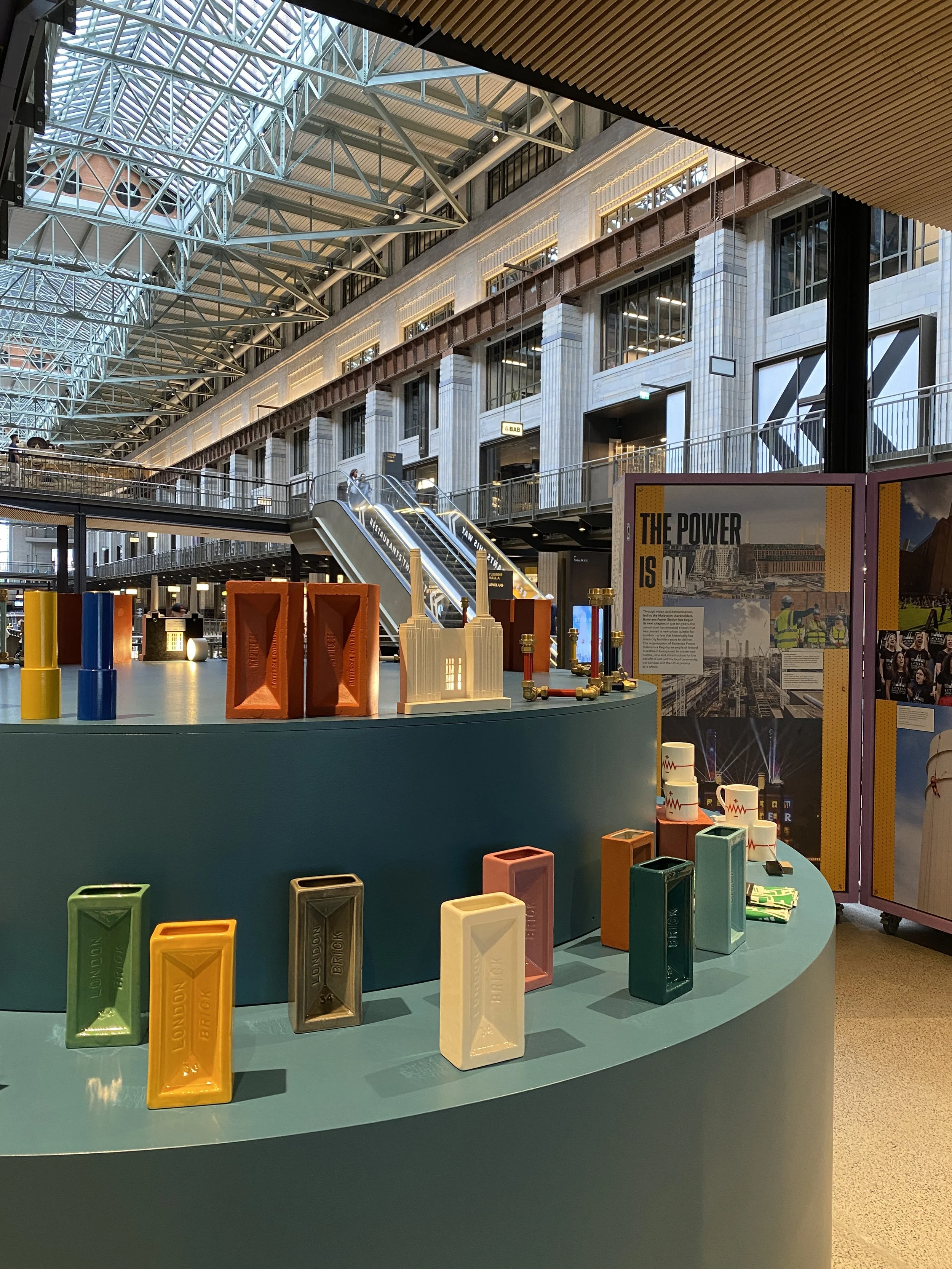 Exhibit display with colorful rectangular boxes and a large poster about power, set in a spacious, modern building with glass ceiling and multiple levels.