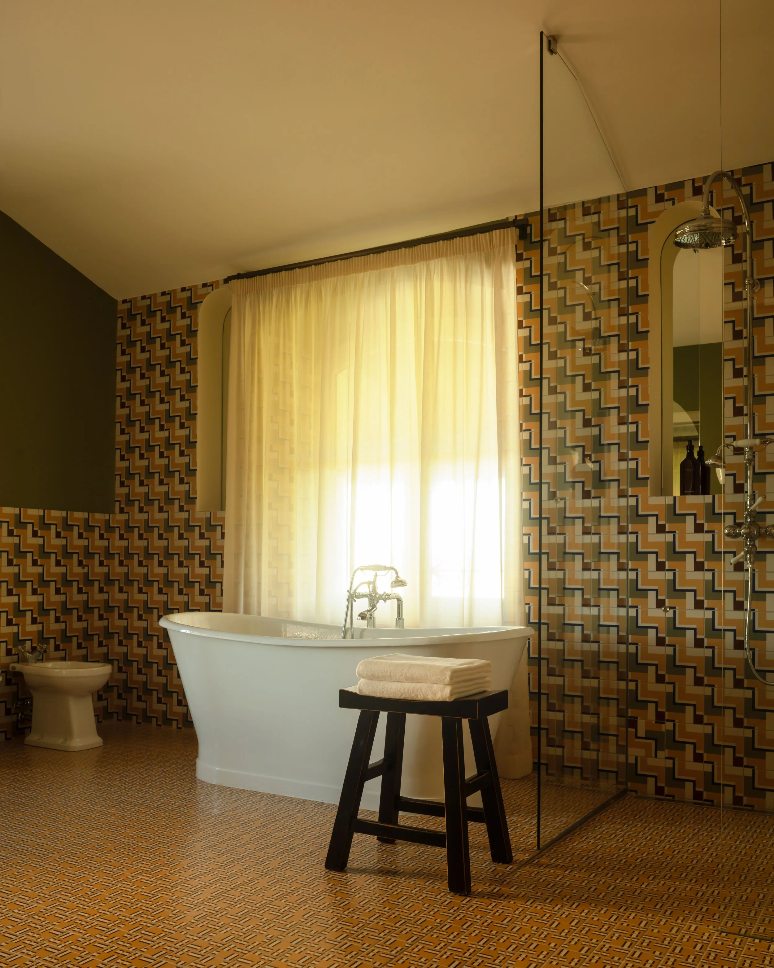 Bathroom with vintage geometric patterned wallpaper, a white bathtub, a small black stool with folded towels, a bidet, a shower with a glass partition, and a window with sheer curtains.