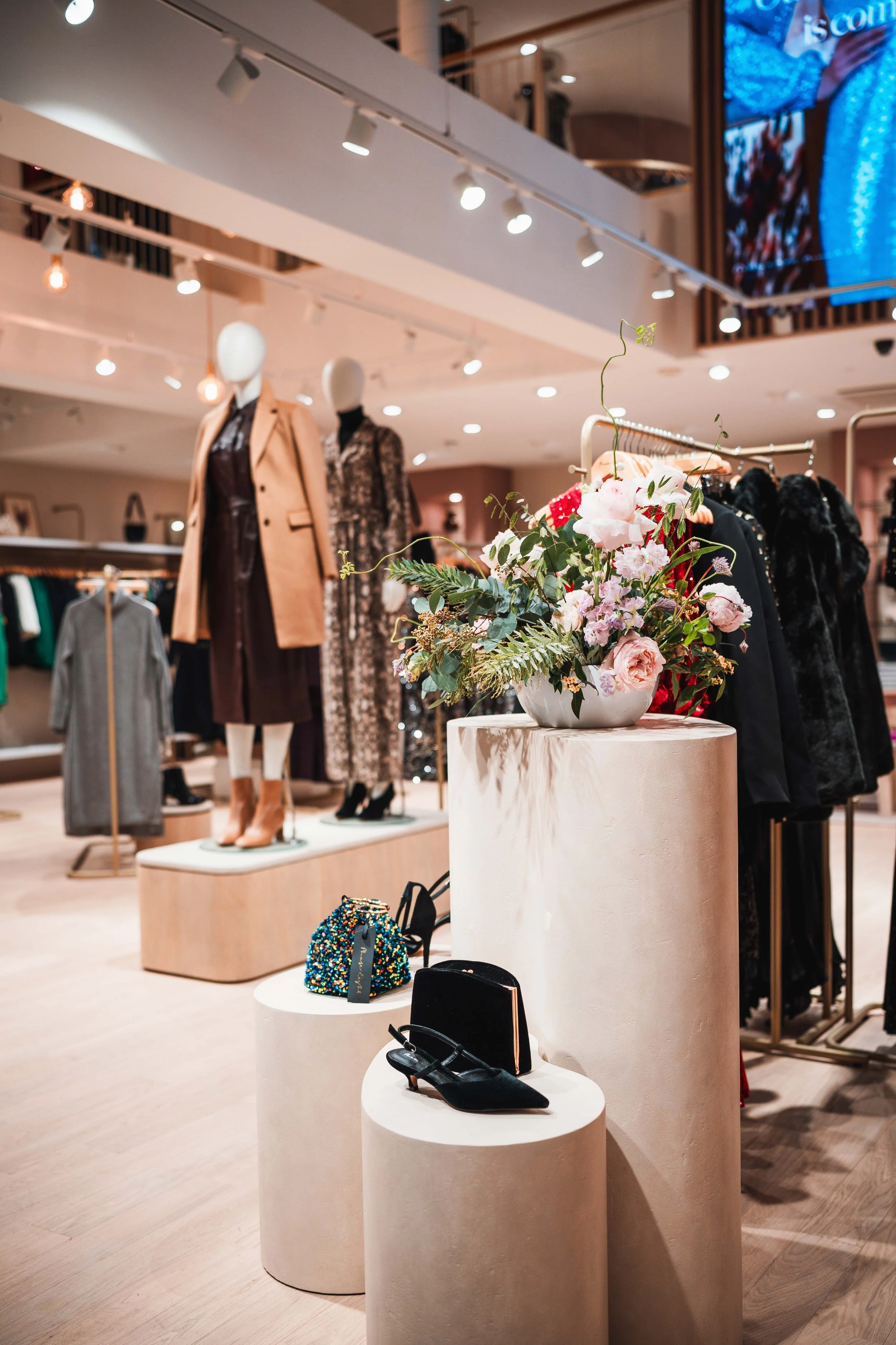 Inside a clothing store displaying mannequins dressed in fashionable outfits, with accessories like shoes and bags on pedestals, and a vase of pink and white flowers on a tall cylindrical pedestal.