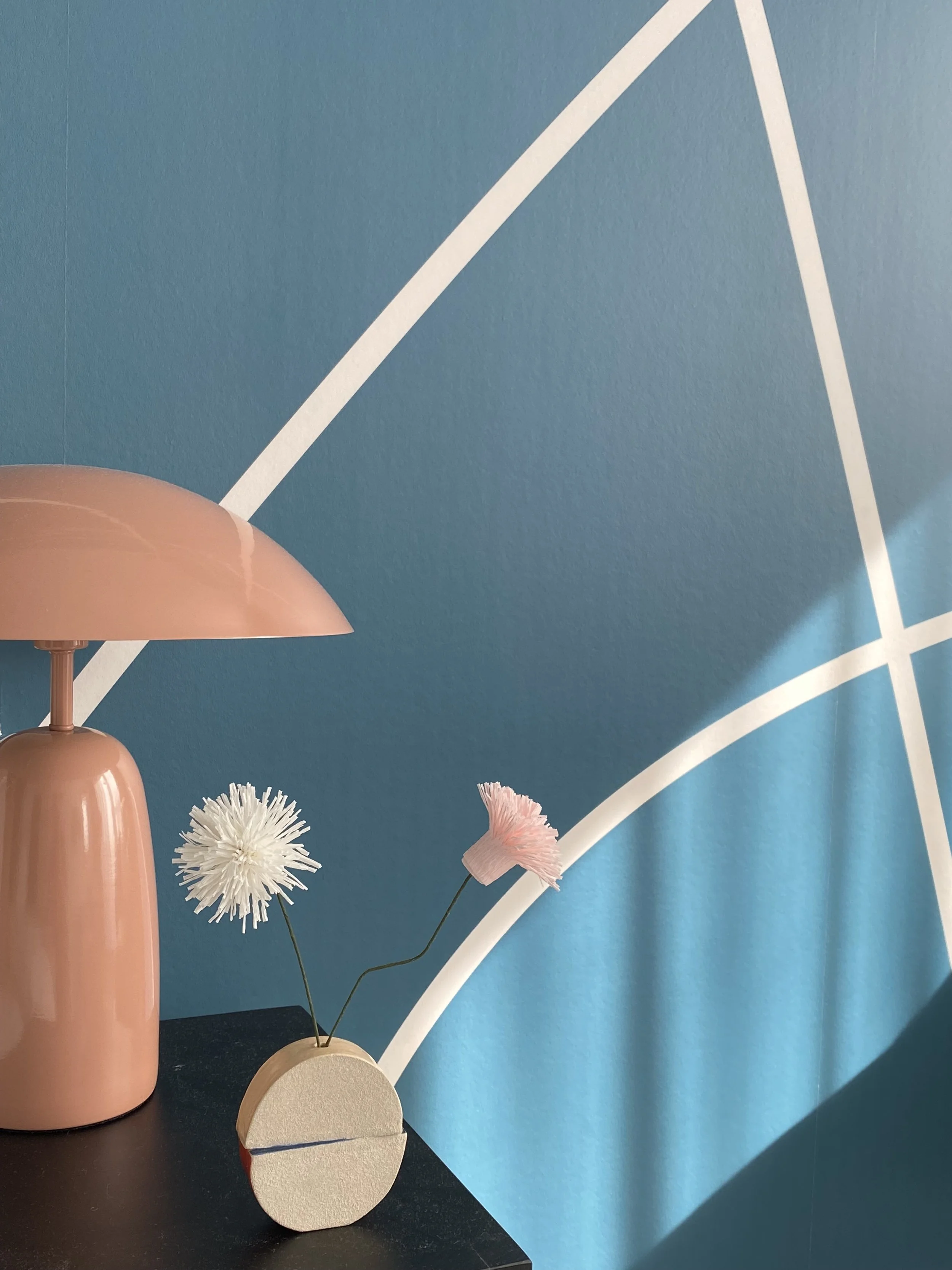A pink table lamp, a decorative vase with pink and white artificial flowers, and a black surface against a blue wall with white geometric lines.