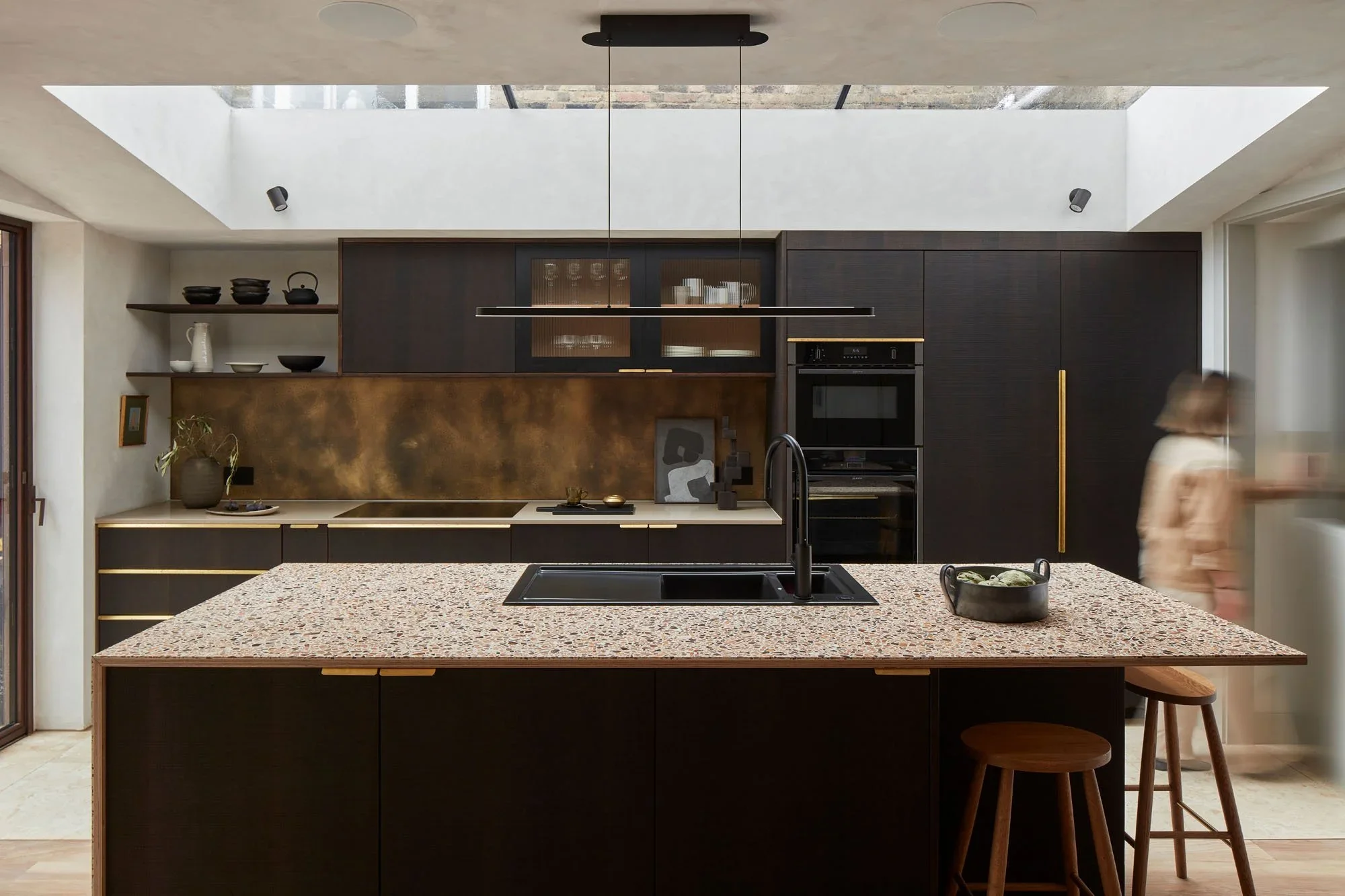 Modern kitchen with a large island counter, black cabinetry, open shelving with bowls and vases, and a skylight overhead. A woman is blurred as she walks past.