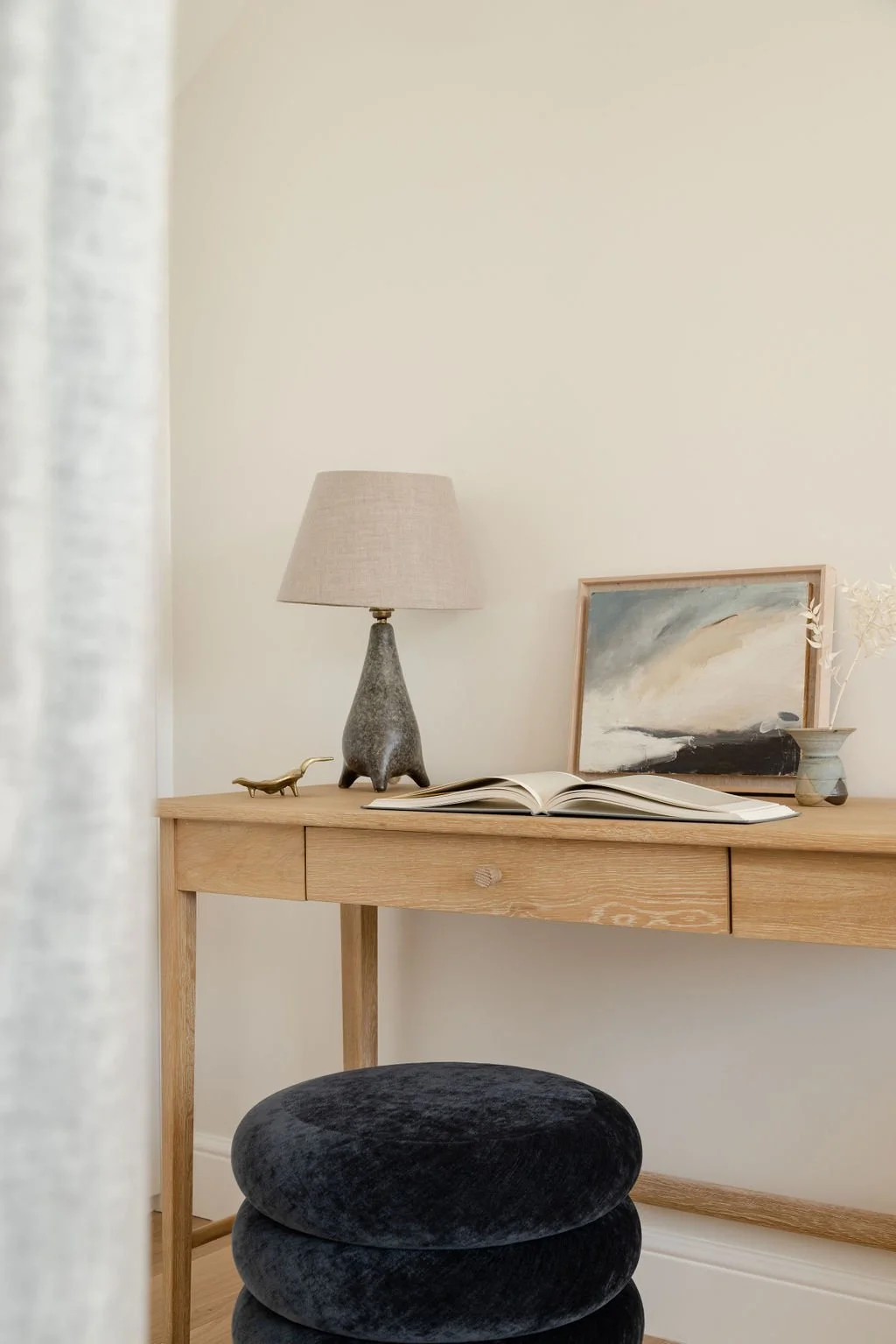 A wooden desk with a table lamp, an open book, a small art painting, a decorative vase with dried flowers, and a small metallic animal figure. In front of the desk is a black velvet cushioned stool. Part of a sheer curtain is visible on the left side