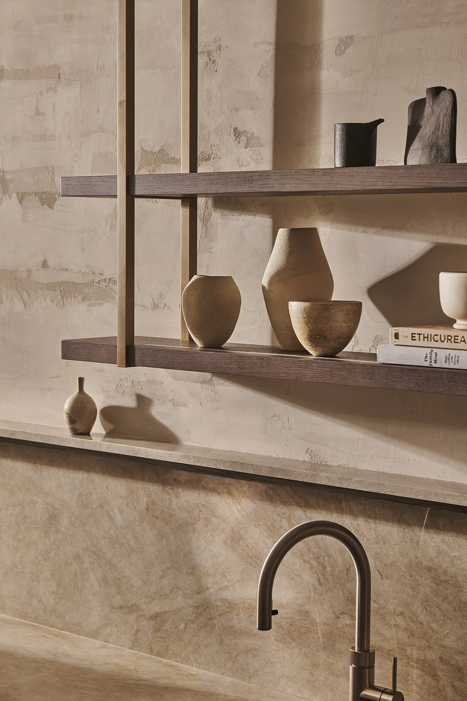 Close-up of a kitchen shelf with various beige and black ceramic vases and a book, with a beige marble backsplash and a metallic kitchen faucet in the foreground.