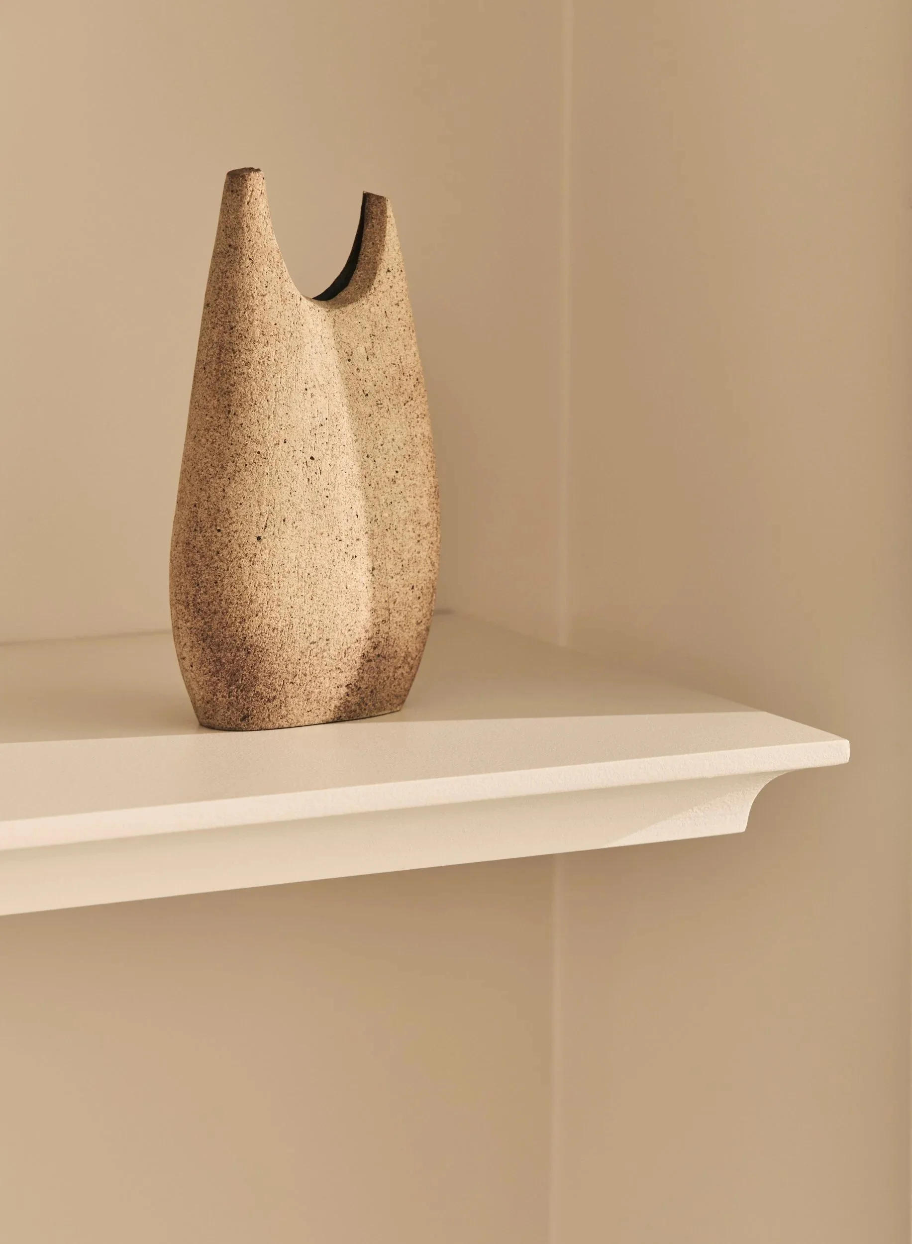 A textured, curved ceramic vase with a slit at the top, placed on a white shelf against a beige wall.