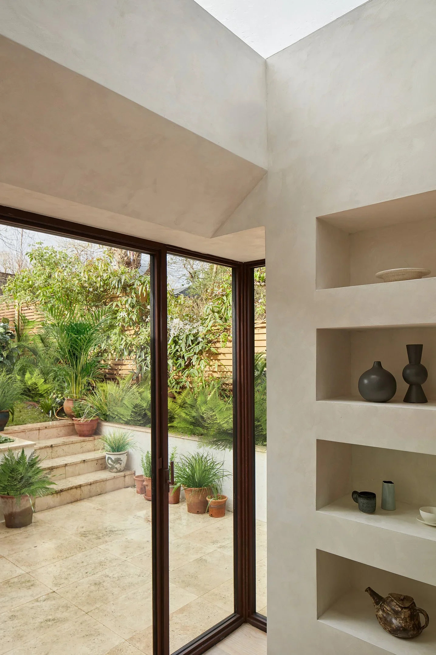 Interior view of a modern home with a beige built-in wall shelf containing vases and bowls, and a glass door leading to a patio with potted plants and greenery outside.
