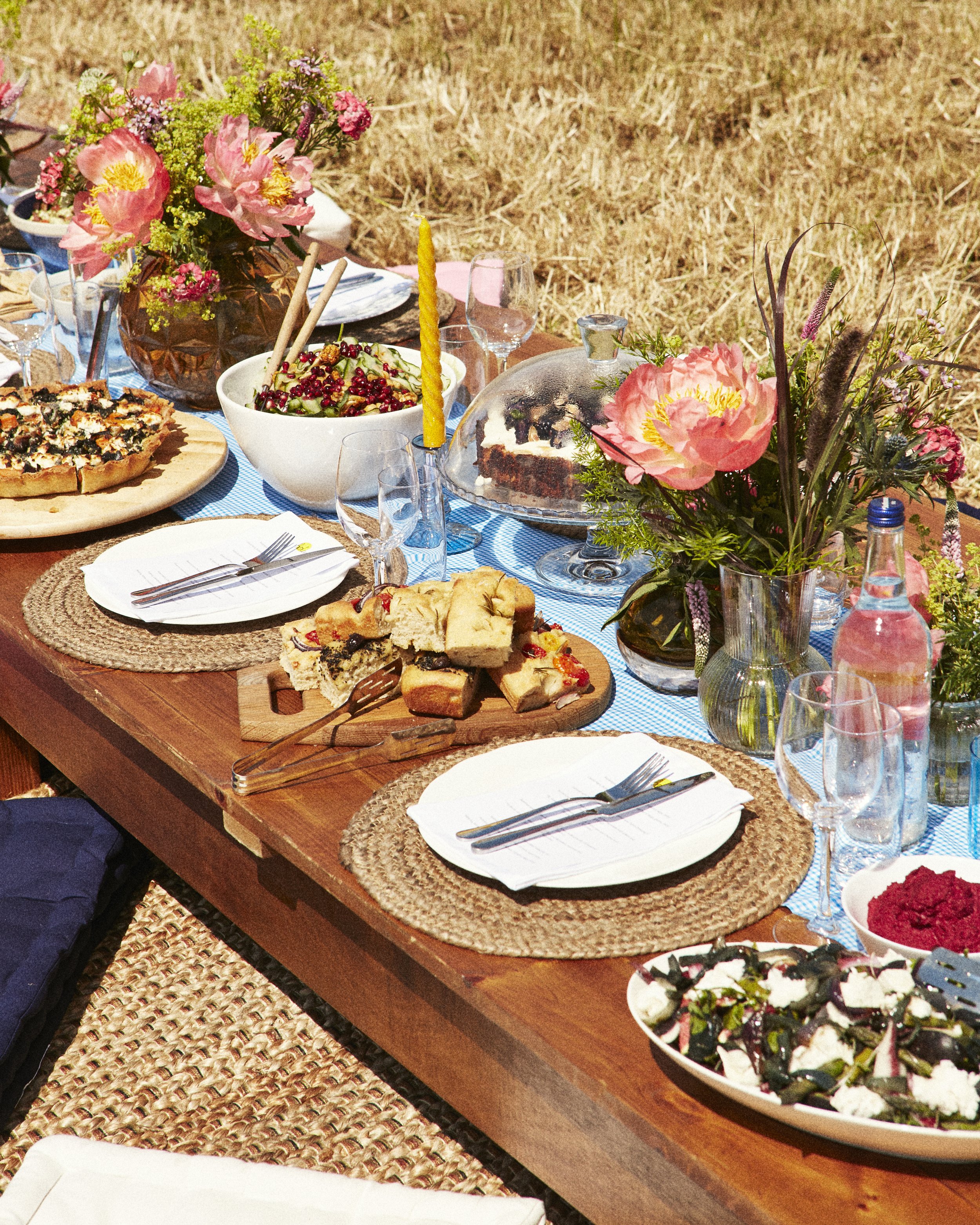 A table set for an outdoor meal with floral arrangements, salads, desserts, a pizza, bread, and drinks, all on a wooden table with wicker placemats and a blue table runner.