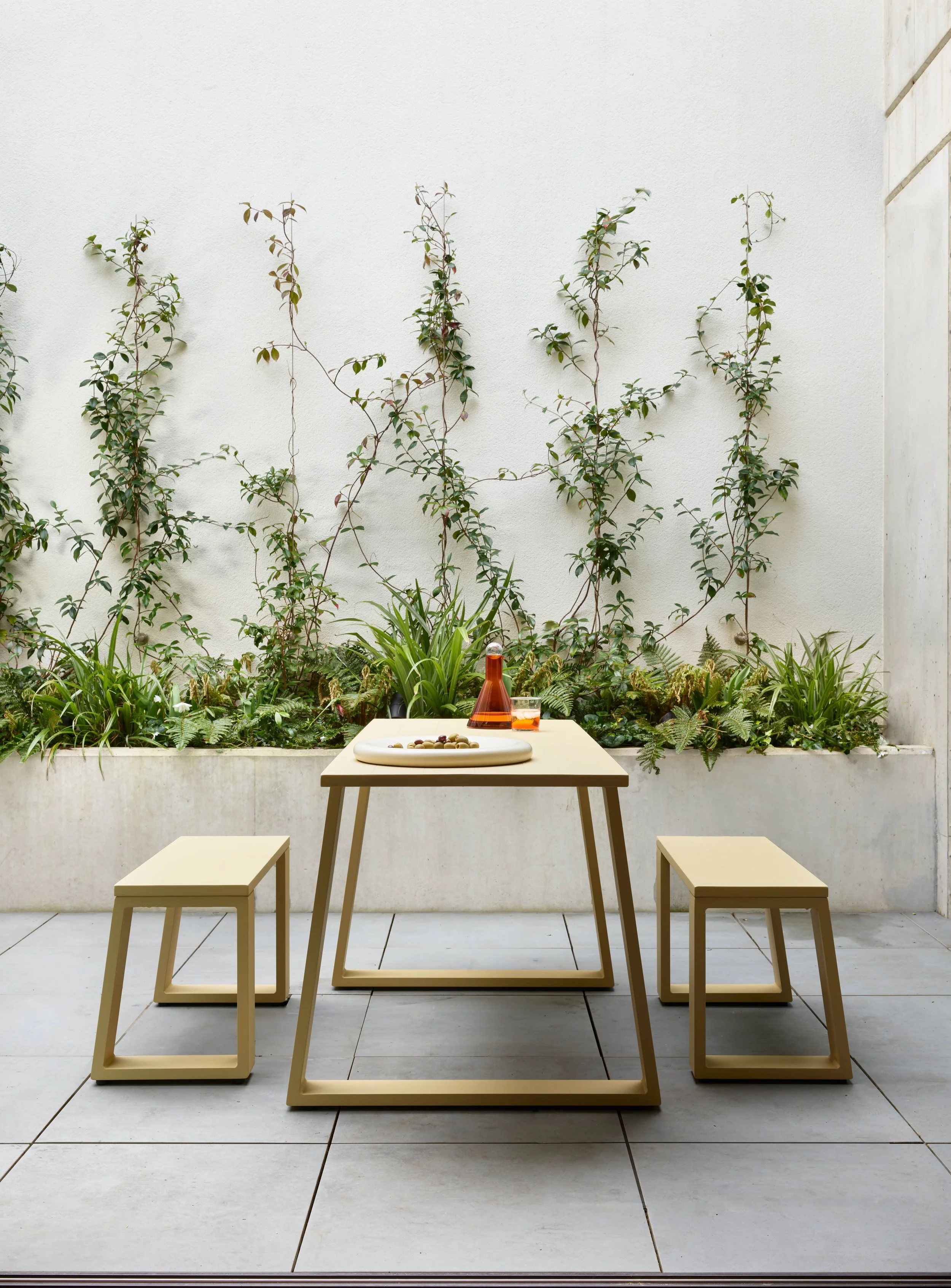 A minimalist outdoor patio with a rectangular table and two matching benches, set against a white wall with climbing plants and greenery.