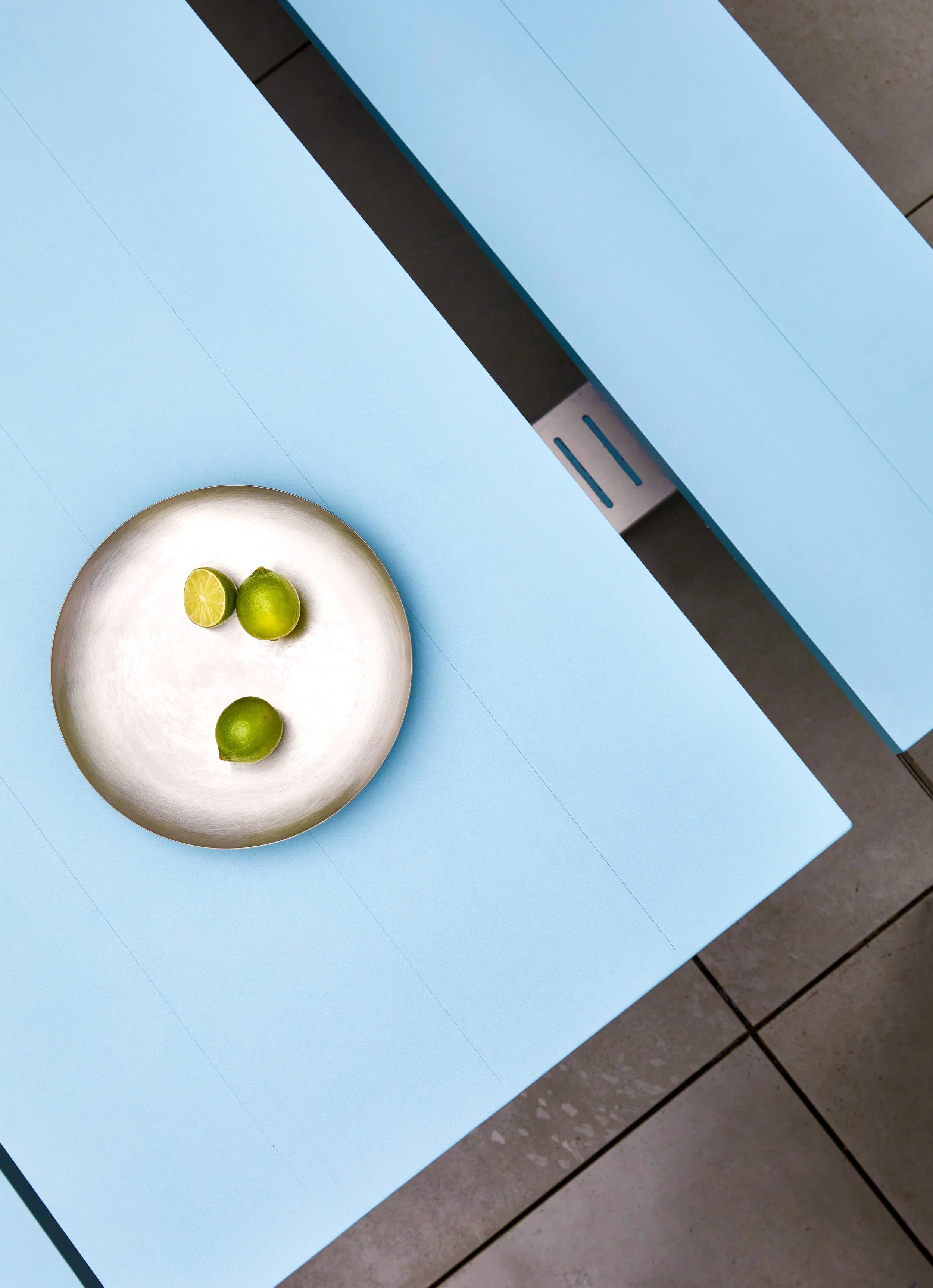 A silver plate with three limes, one cut in half, on a light blue table with a matching blue chair nearby.