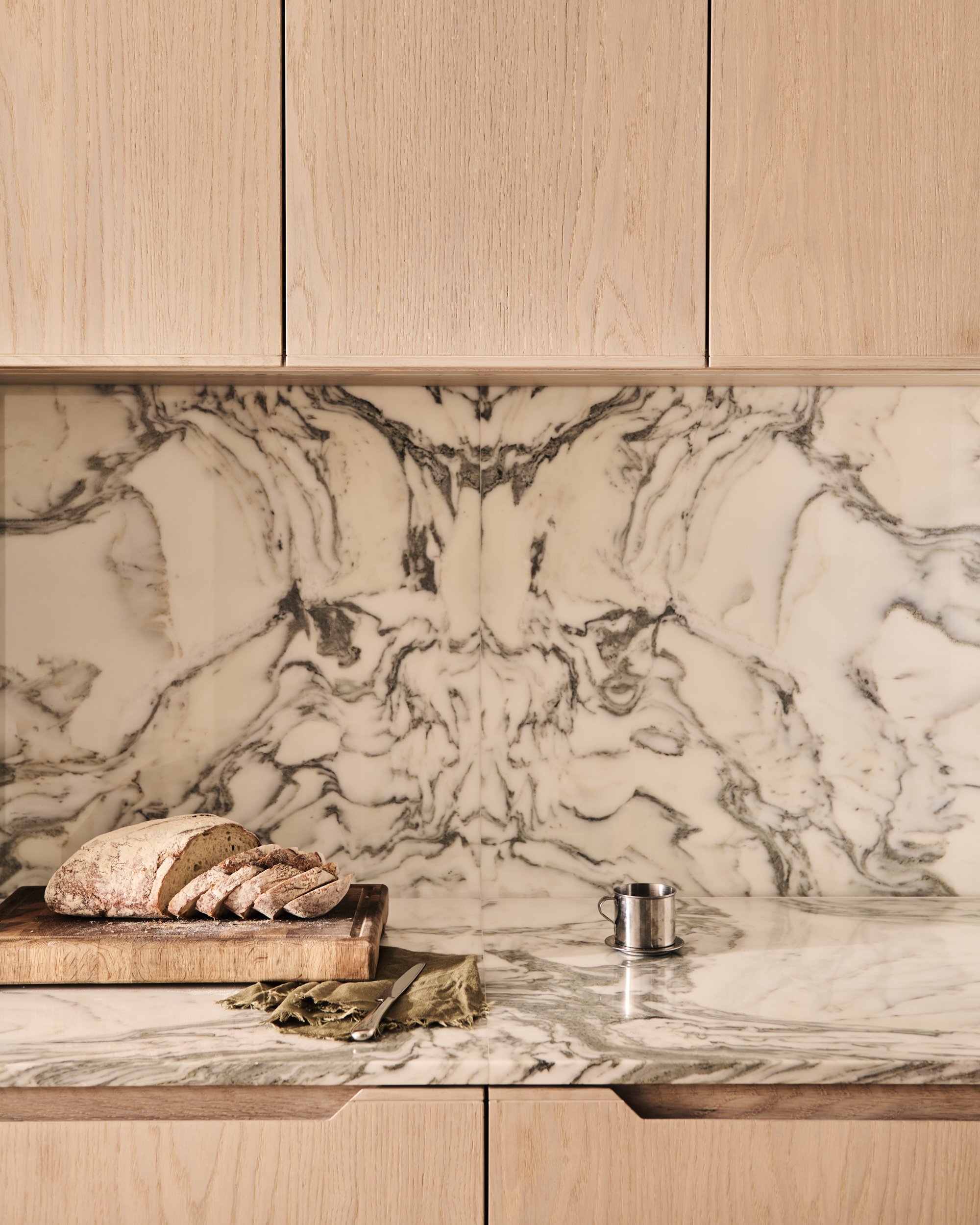 Kitchen countertop with sliced bread on a wooden cutting board, a knife, a metal cup, and a green cloth.