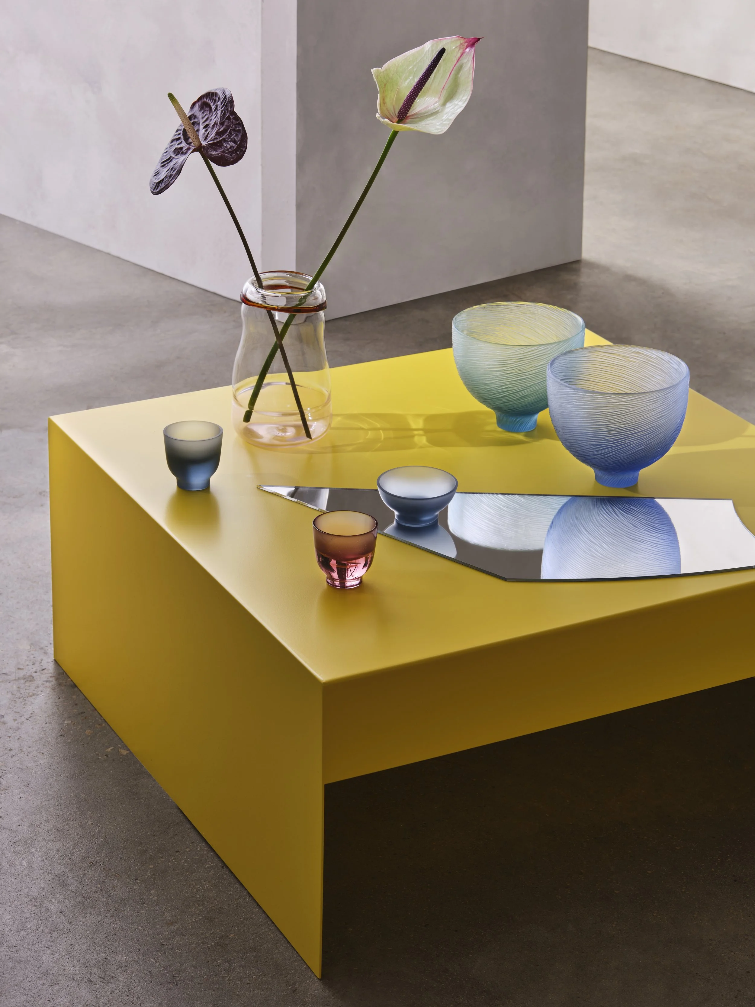 Decorative glassware on a yellow table, including a pink vase with flowers and various ceramic bowls and cups.