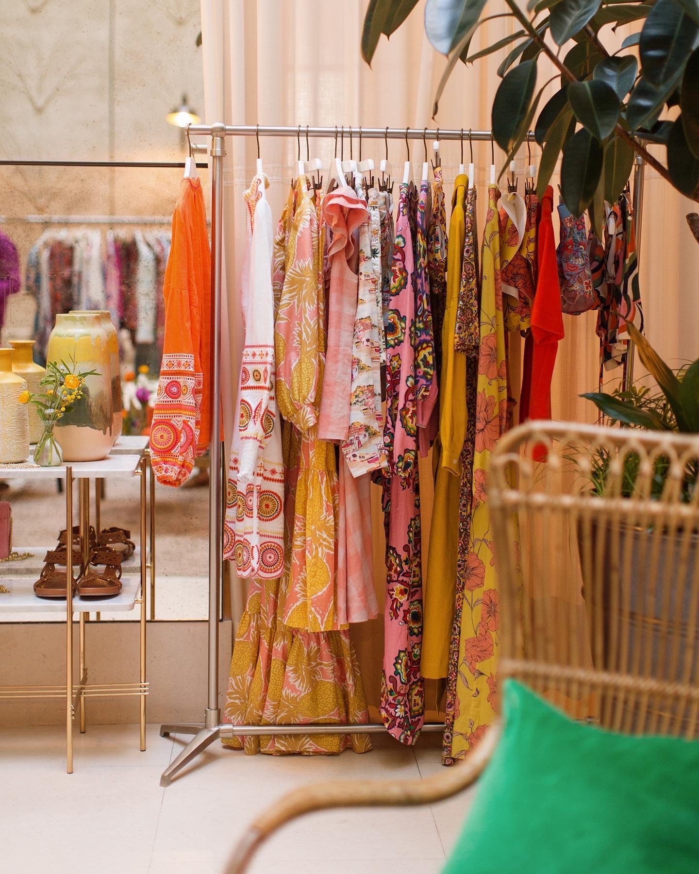 A clothing rack with colorful dresses and garments, a small table with decorative vases and shoes, and indoor plants, in a boutique store.
