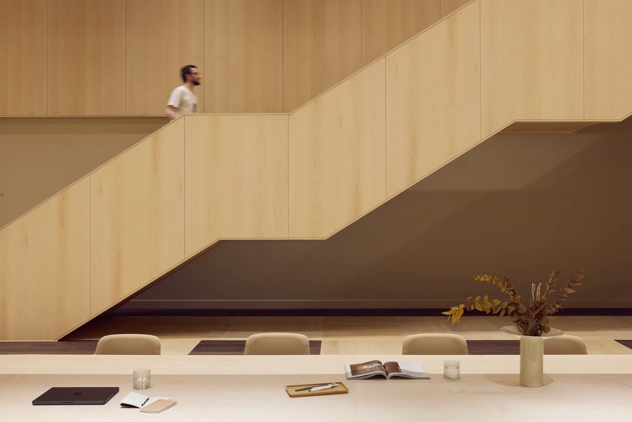 Interior view of a modern space with a light-colored wooden staircase, beige chairs, a long table with a laptop, open book, notebooks, pens, a vase with dried flowers, and glasses of water.