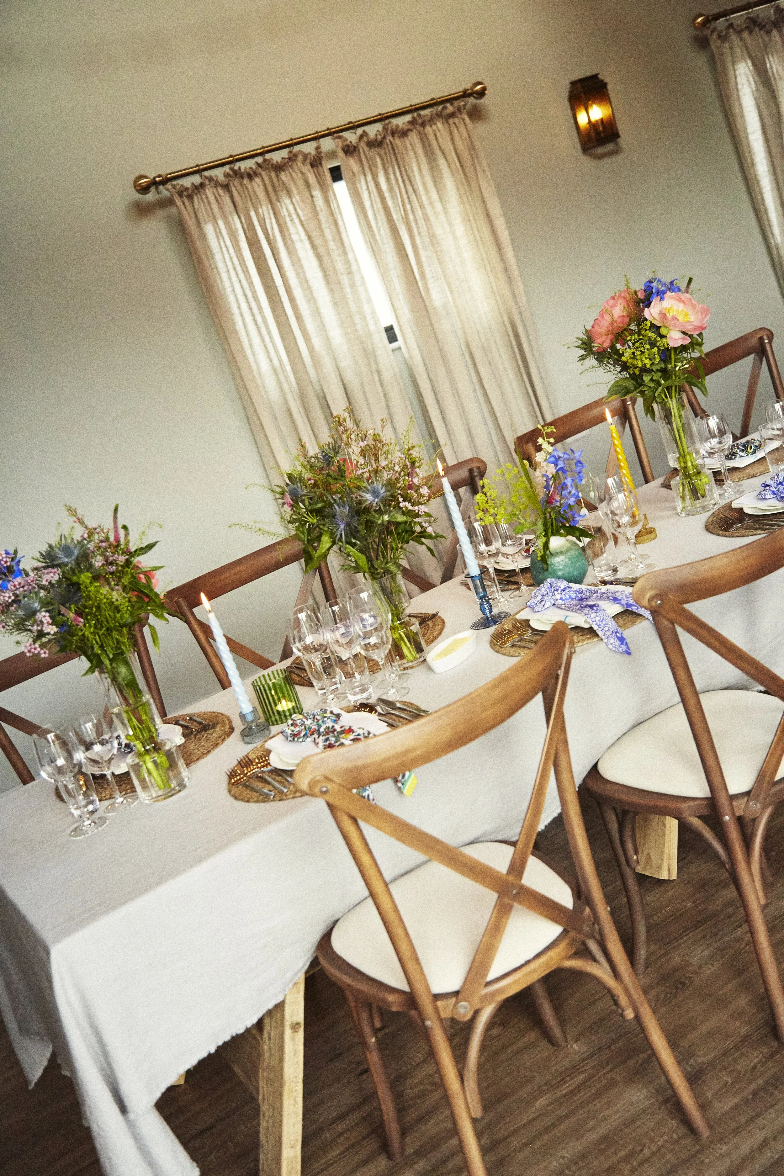 A dining table decorated with flowers, candles, and place settings in a cozy room with beige curtains and wooden floors.