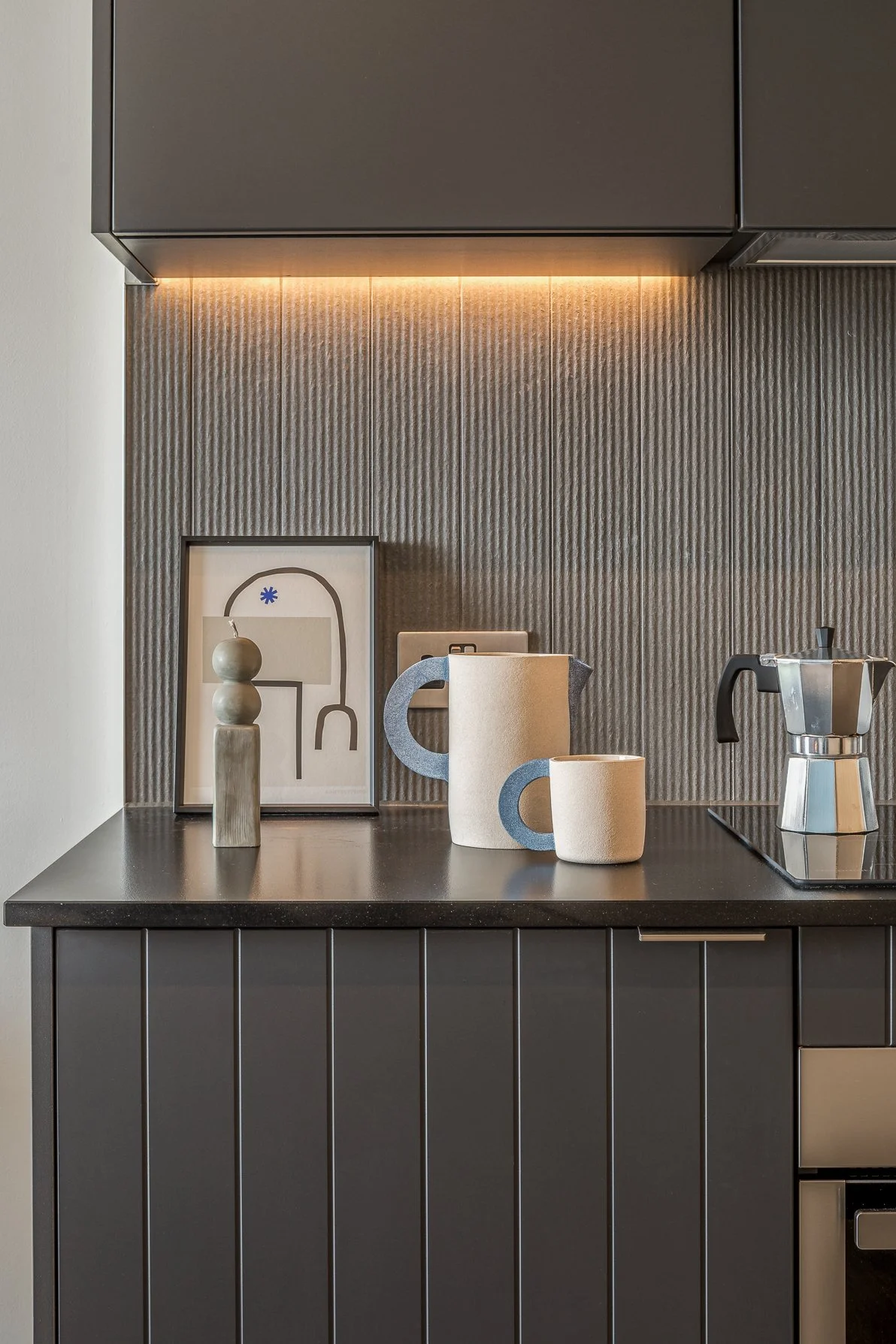 Minimalist kitchen countertop with beige and blue ceramic mugs, framed abstract art, a small decorative statue, and a silver moka pot on the stove.