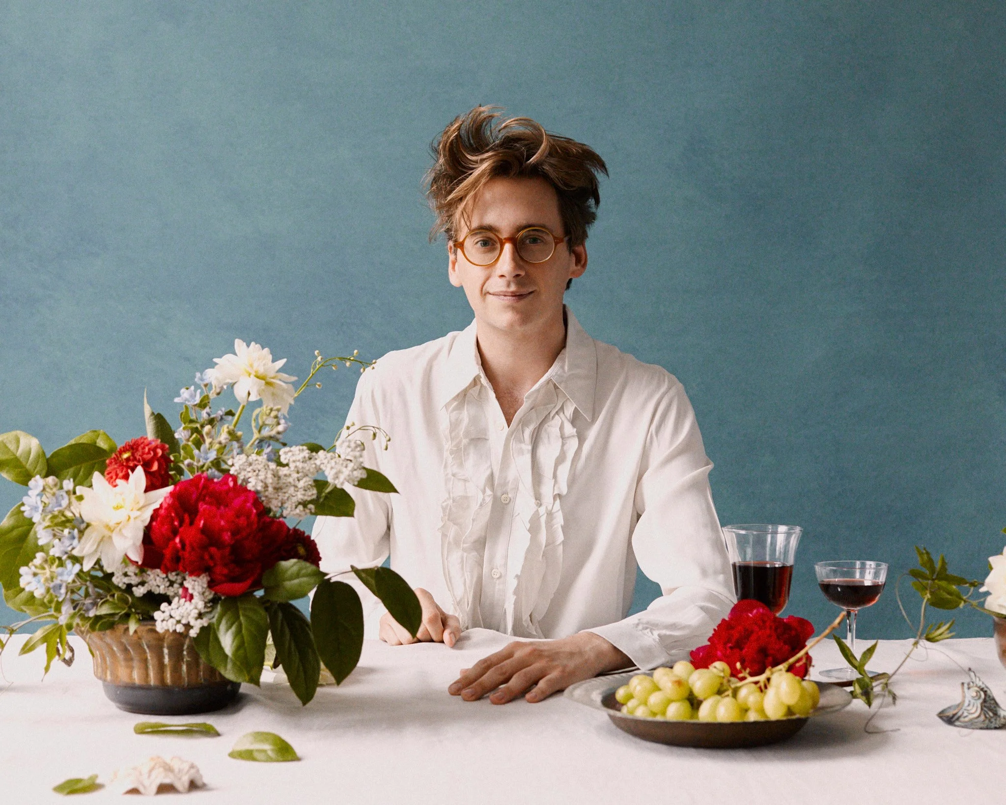 A young man with glasses and wavy hair sitting at a table decorated with flowers, grapes, and glasses of wine, against a teal background.