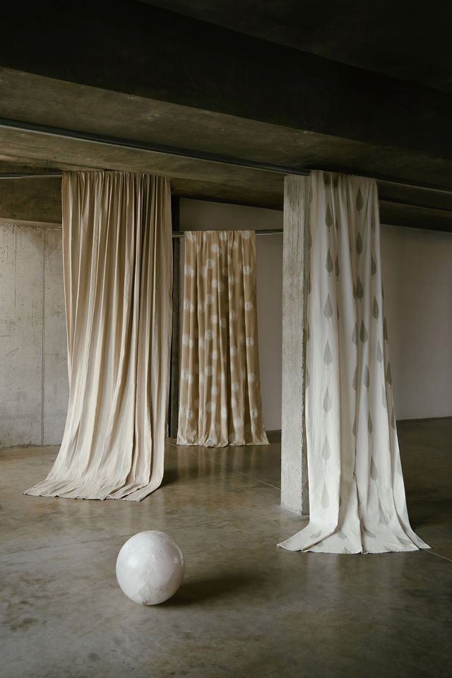 Three elegant curtains hanging from a concrete ceiling in an industrial-style space, with a marble sphere on the floor in front of them.