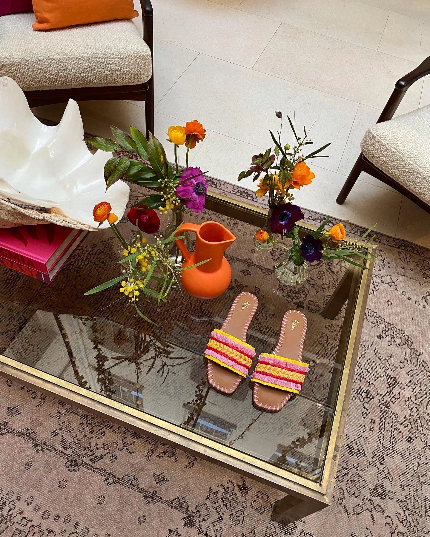 A glass-top coffee table with pink, yellow, and purple slippers, a vase of colorful flowers, an orange vase, and decorative objects. Surrounding furniture includes upholstered chairs and a patterned rug.