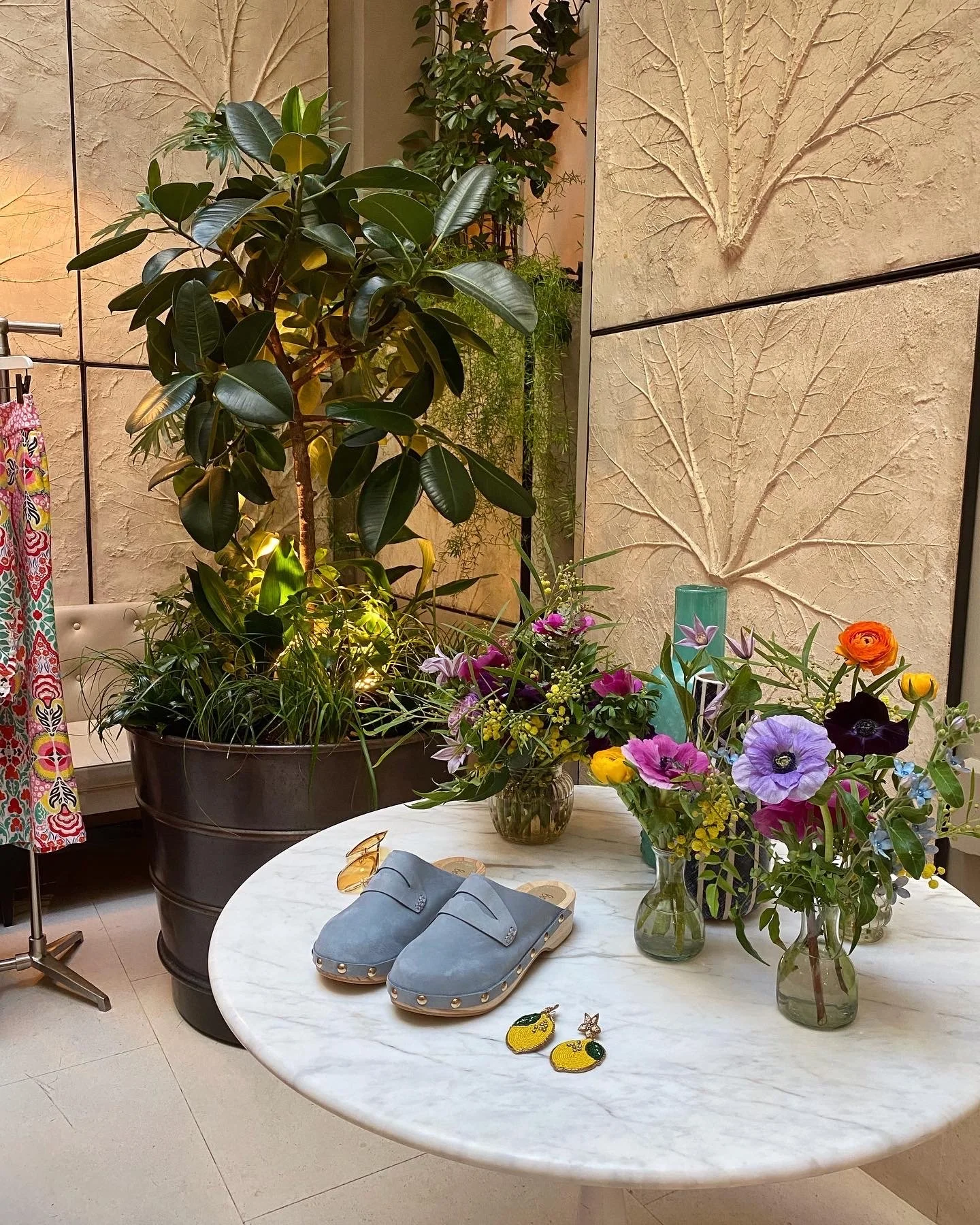 A round marble table with three vases of colorful flowers, a pair of blue slippers, and a pair of yellow and green embroidered earrings. Large potted plants and decorative textured walls are in the background.