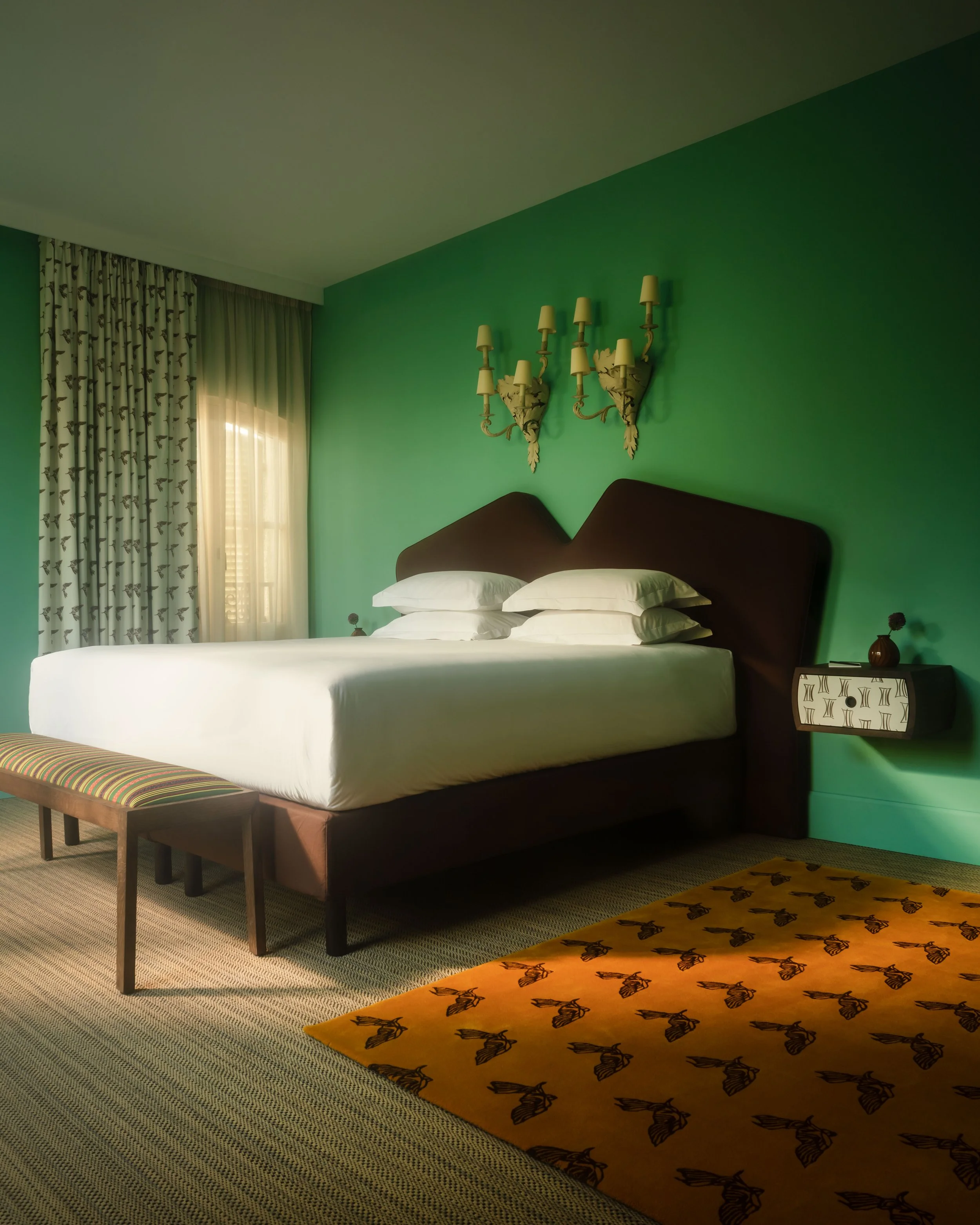 Hotel room with a white bed, dark headboard, green wall, wall-mounted nightstand, window with curtains, and a yellow rug with black bird pattern.