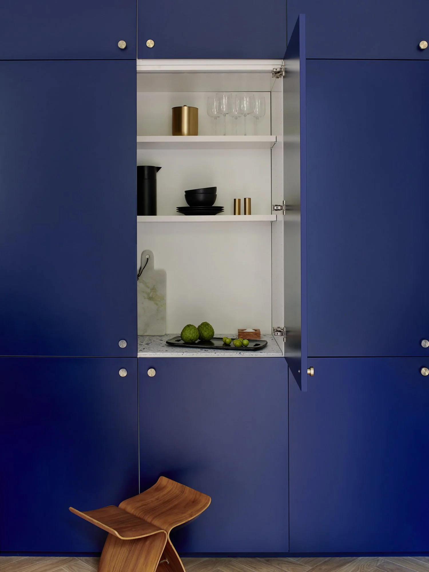 Open blue cabinet with white interior, containing glassware, black and gold tableware, and artichokes on a tray, with a small wooden stool in front.